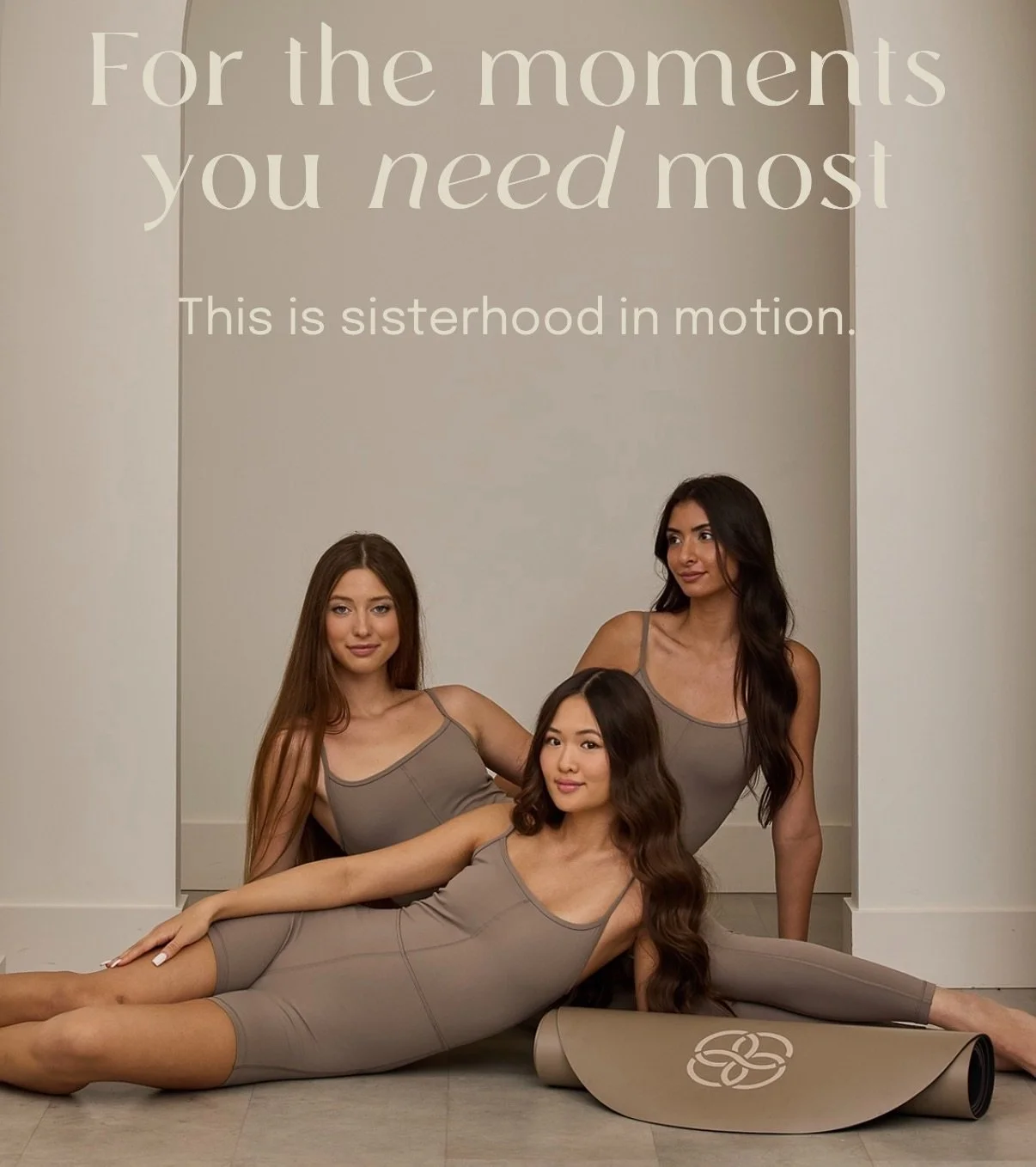 Three women in matching yoga outfits practicing yoga in front of a wall with motivational text. One woman is lying on the floor with yoga mat unrolled, and two women are sitting and kneeling behind her.