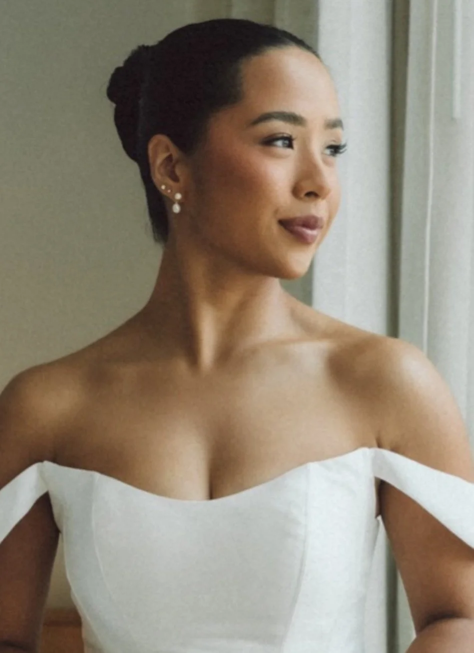 A woman with dark hair styled in an updo, wearing pearl earrings, looking out a window with a soft expression, dressed in an off-shoulder white dress.