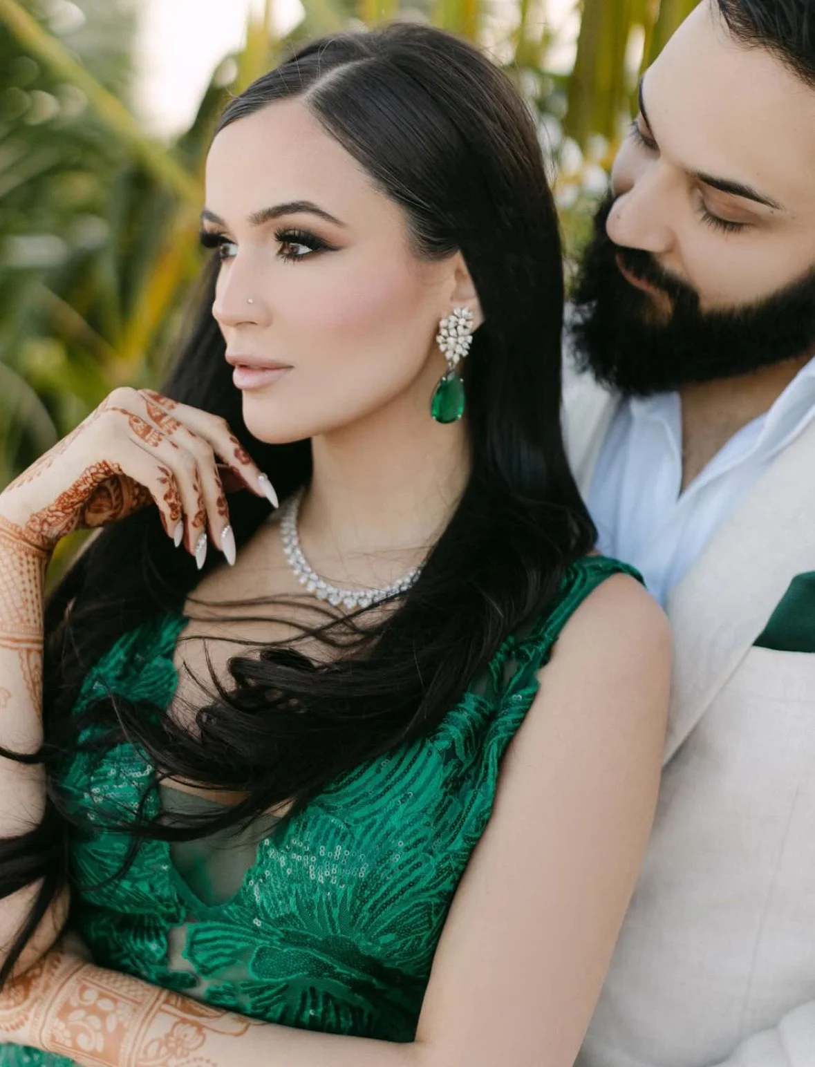 A woman with long black hair, green earrings, an emerald dress, and henna on her hand, while a bearded man in a white suit stands close to her.