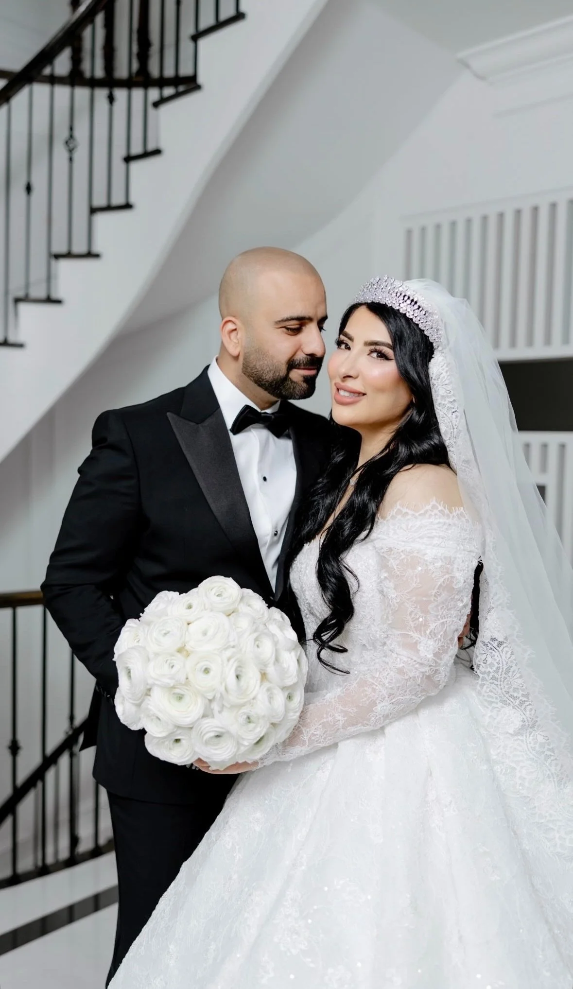 A bride and groom pose together indoors, with the bride holding a bouquet of white roses. The groom wears a black tuxedo and bow tie; the bride wears a white lace wedding gown with a veil and tiara. They are standing near a staircase.