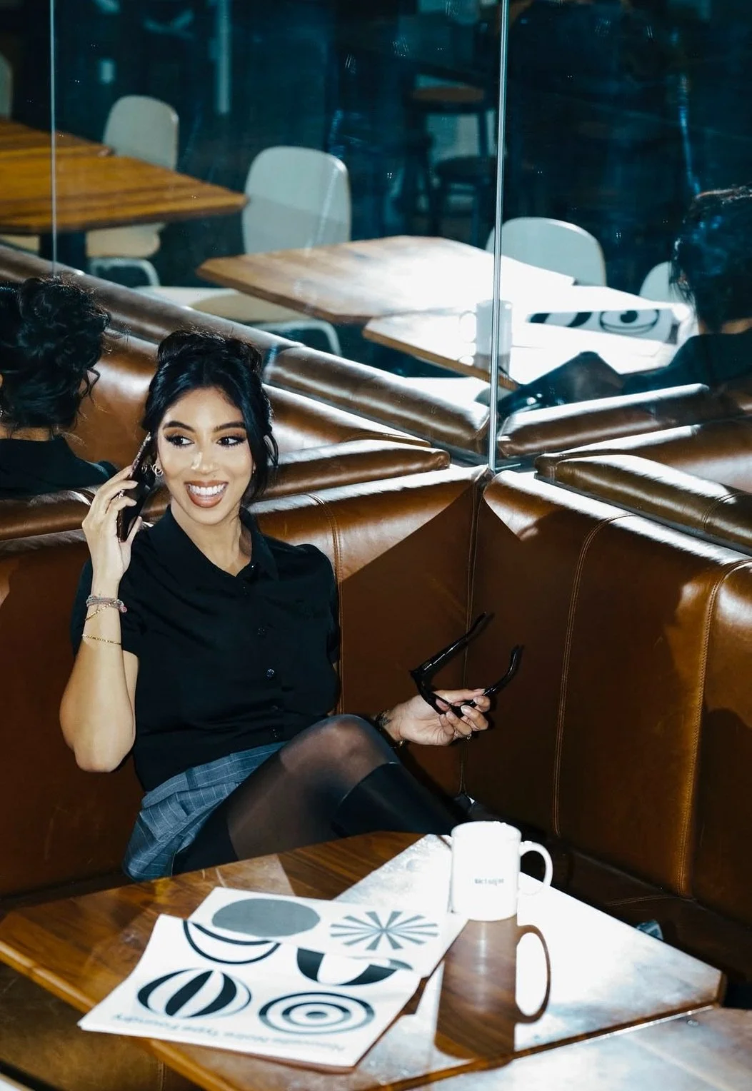 A woman sitting on a brown leather booth, talking on a cellphone, holding sunglasses, with a white mug and patterned paper on the table, in a restaurant with glass walls and empty tables in the background.
