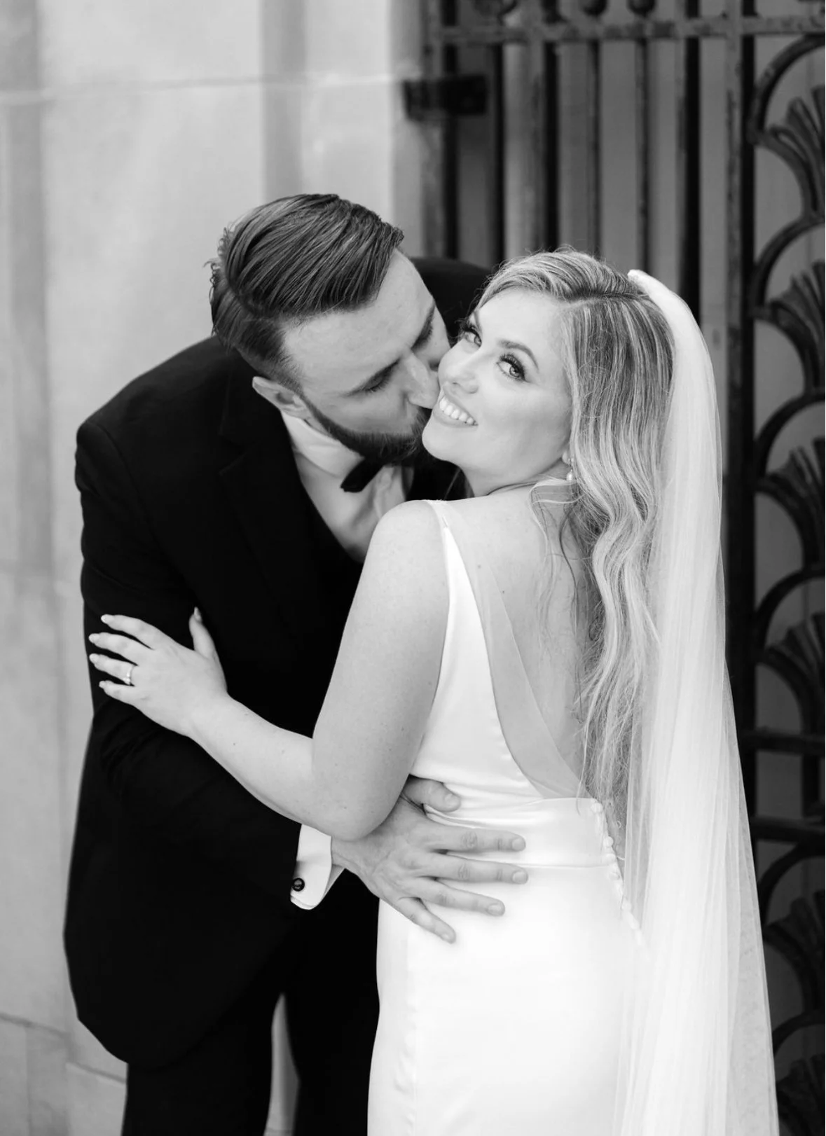 A wedding couple in formal attire embracing and smiling, with the groom kissing the bride on the cheek, in a black and white photo.