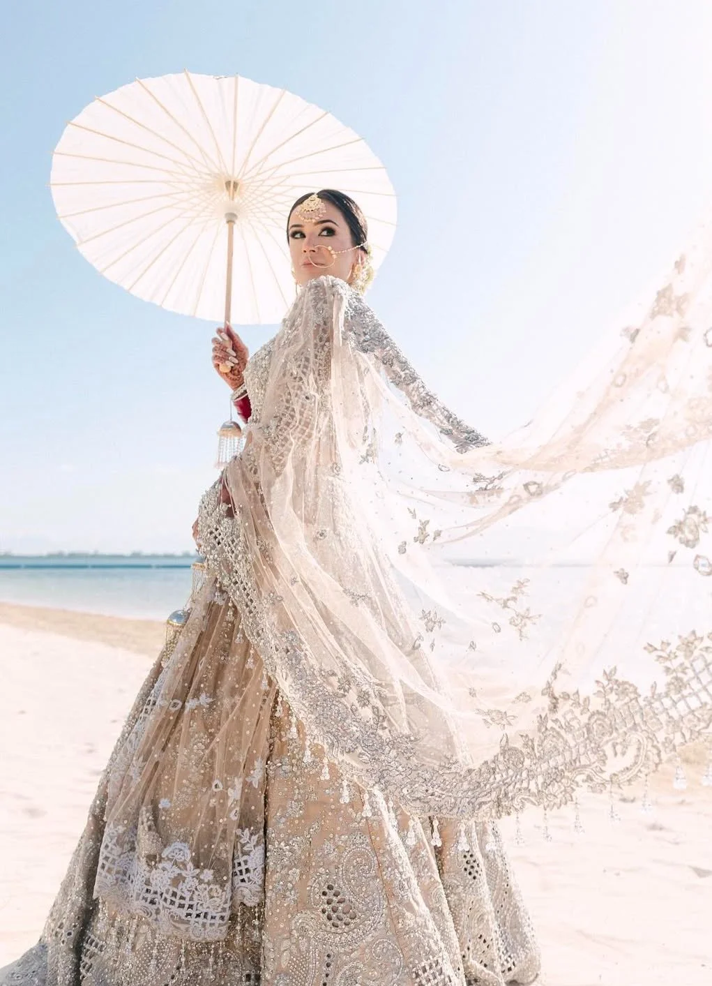 A woman wearing a traditional ornate wedding dress holding a white parasol on a sandy beach with the ocean in the background.
