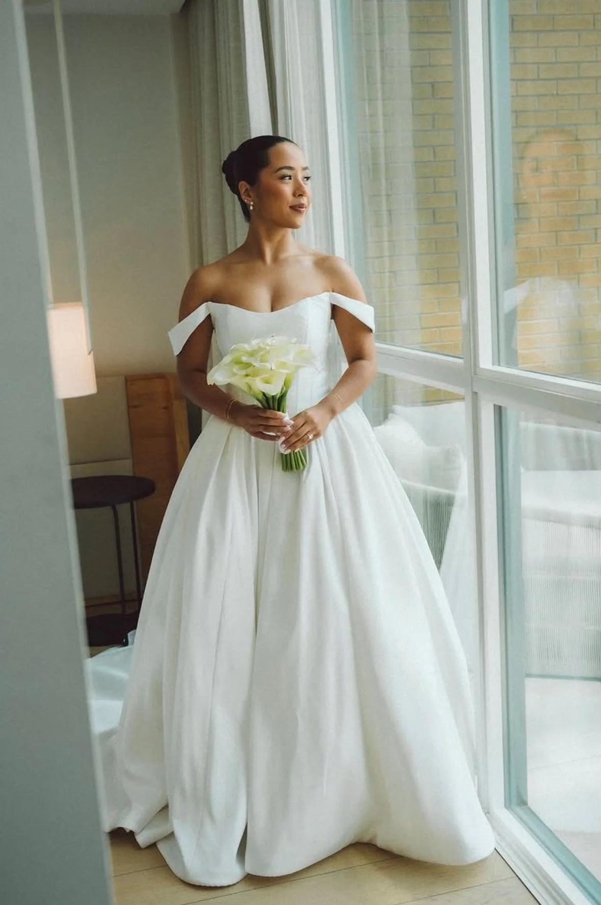 A bride in a white wedding dress holding a bouquet of Calla lilies, standing by a large window and looking outside.