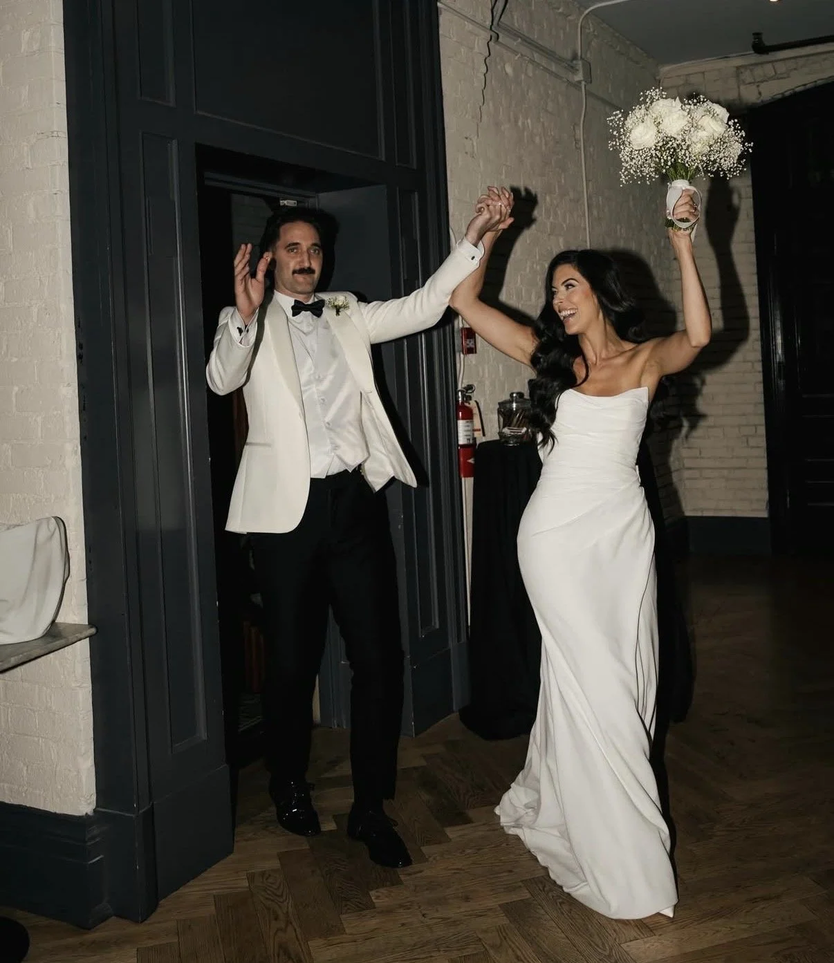 A groom and bride dancing at their wedding reception, holding hands and smiling, with the bride holding a bouquet of flowers.