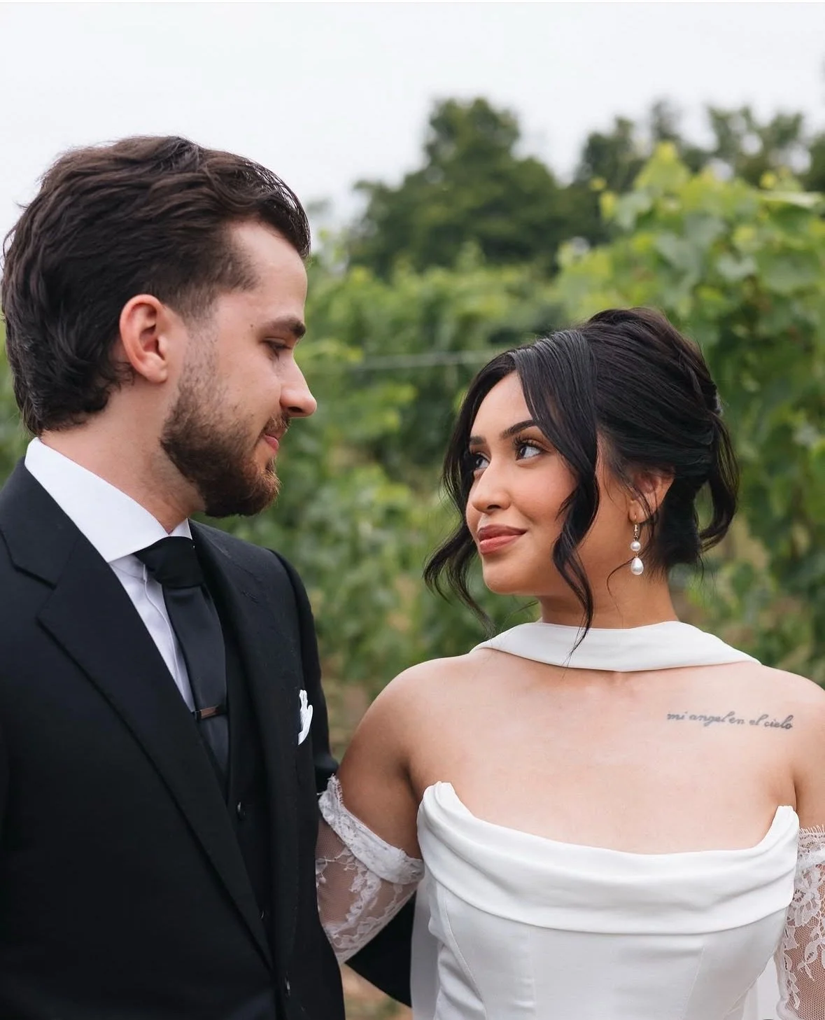 A man and woman standing outside during a wedding, looking at each other. The man is wearing a black suit with a white shirt and black tie. The woman is wearing a white wedding dress with lace sleeves, pearl earrings, and has a tattoo on her shoulder