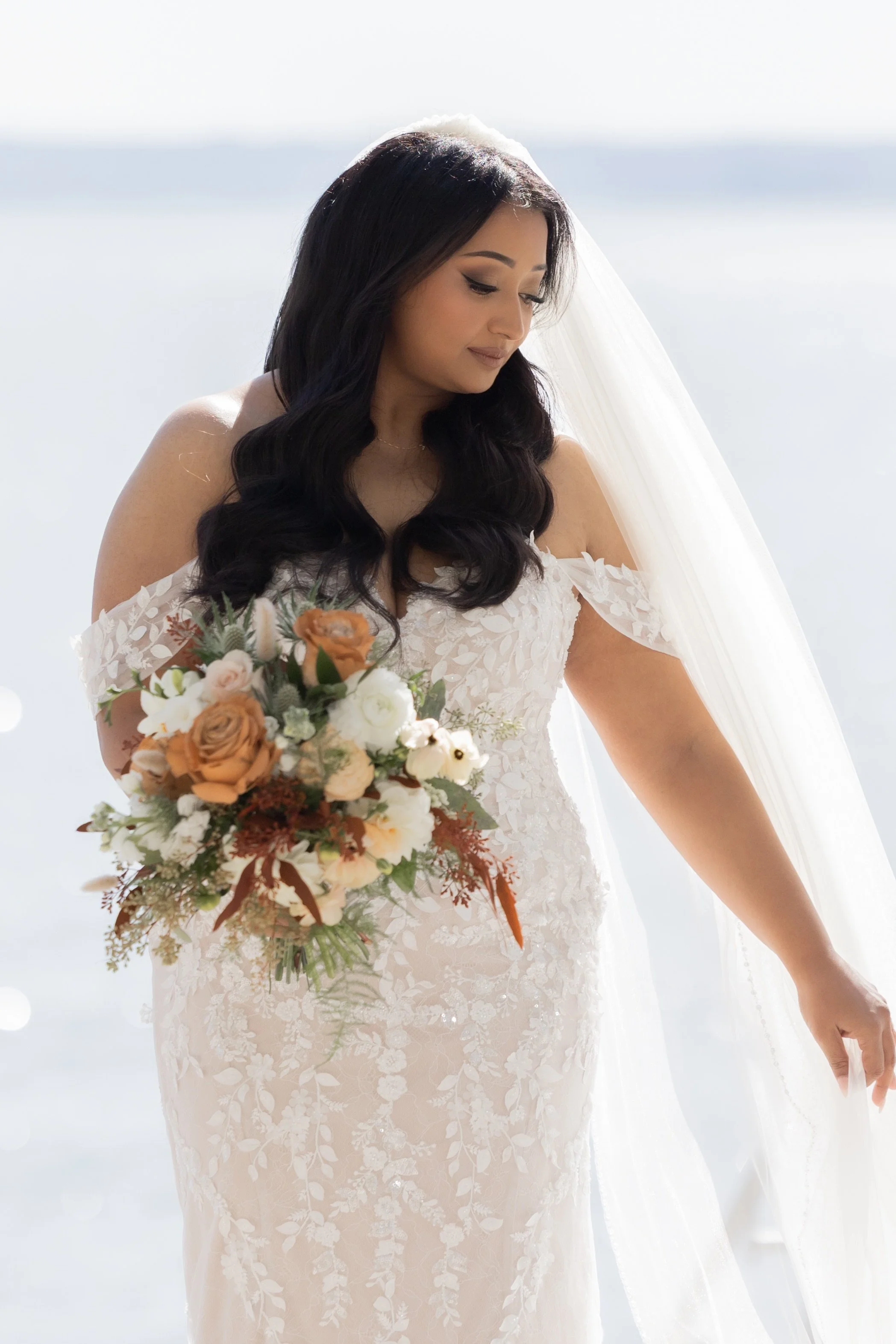 A bride in a white wedding dress holding a bouquet of flowers, standing outdoors in bright sunlight with a blurred body of water in the background.