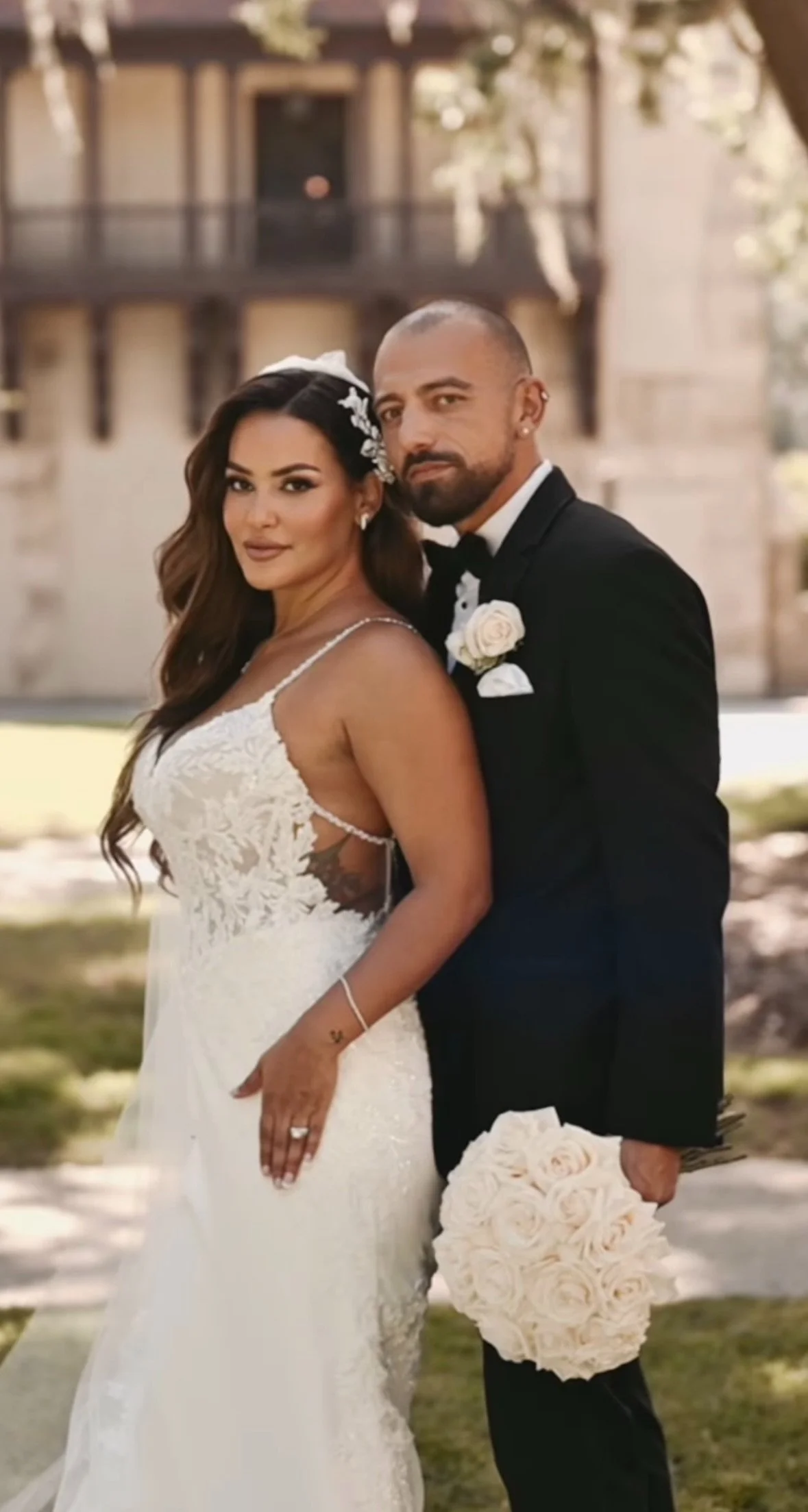 A newlywed couple stands outdoors in front of a building, with the bride wearing a white wedding dress and the groom in a black tuxedo, holding a bouquet of cream roses.