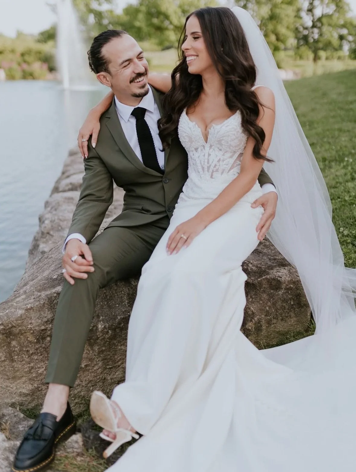 A bride and groom sitting on a rock near a body of water, sharing a joyful moment, with the bride wearing a white wedding gown and veil, and the groom in a green suit and black tie.