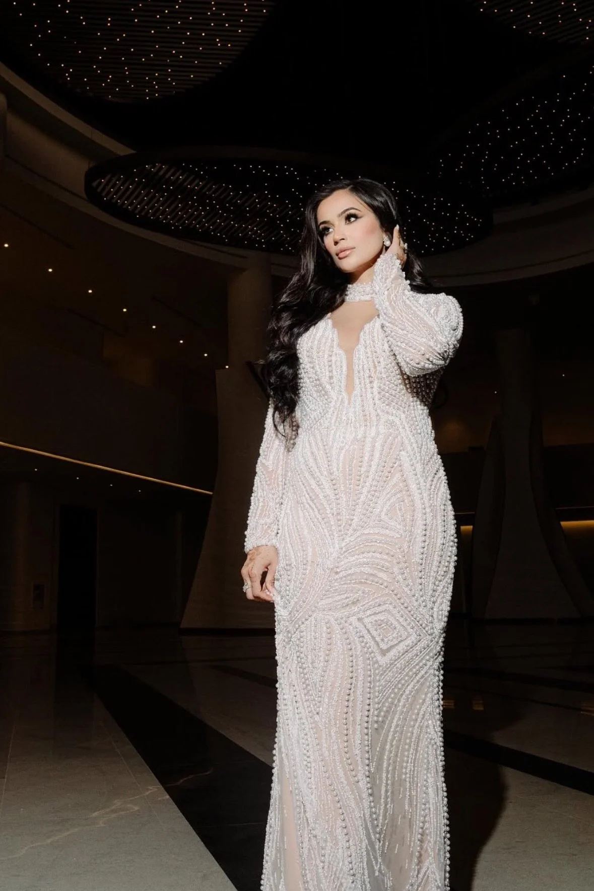 A woman in an elegant, long, white beaded dress standing indoors with a dark background and decorative ceiling lights.