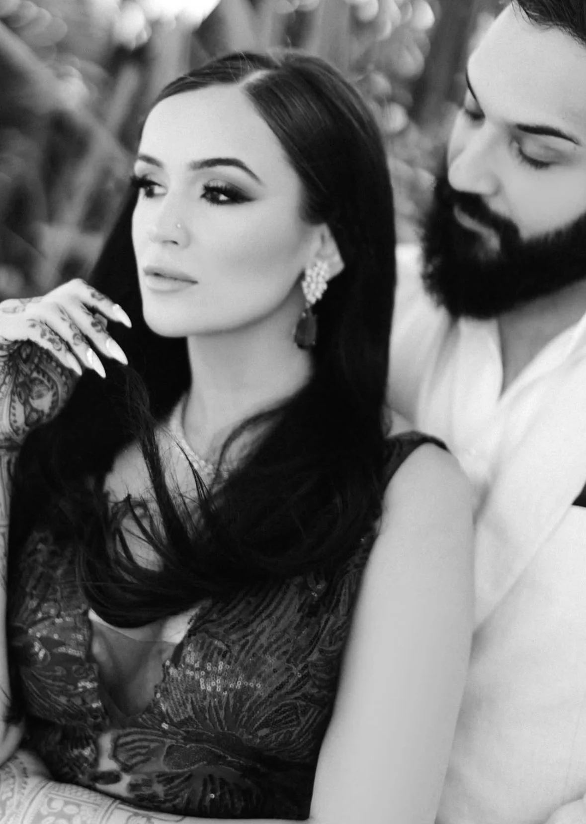 A woman with long dark hair and earrings, and a man with a beard, are in close proximity, with the woman gazing to the side and the man looking at her. The photo is in black and white.