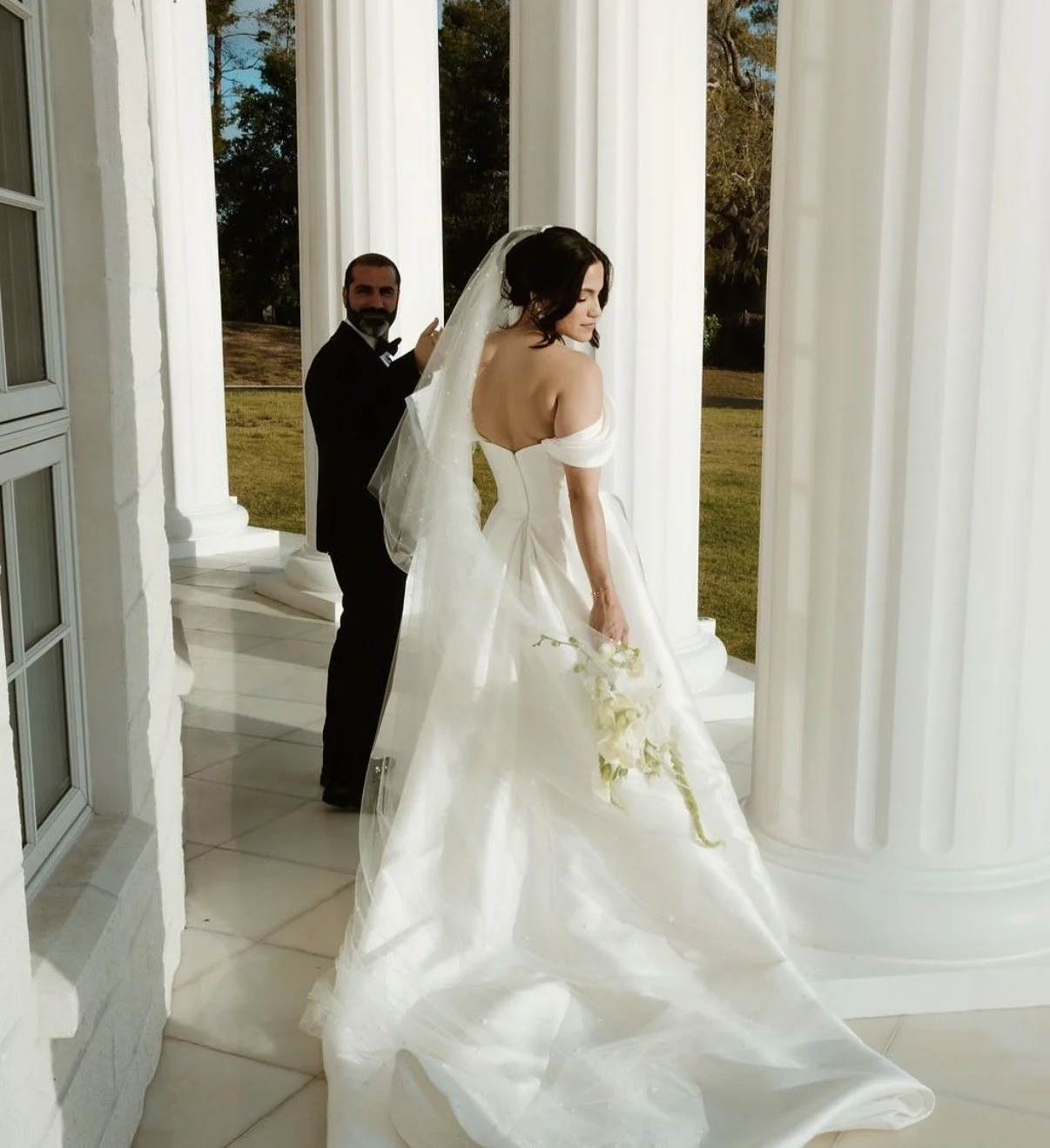 A bride in a white lace wedding dress sitting in a decorated venue, holding a bouquet of white flowers, with a neutral background of large windows and floral arrangements.