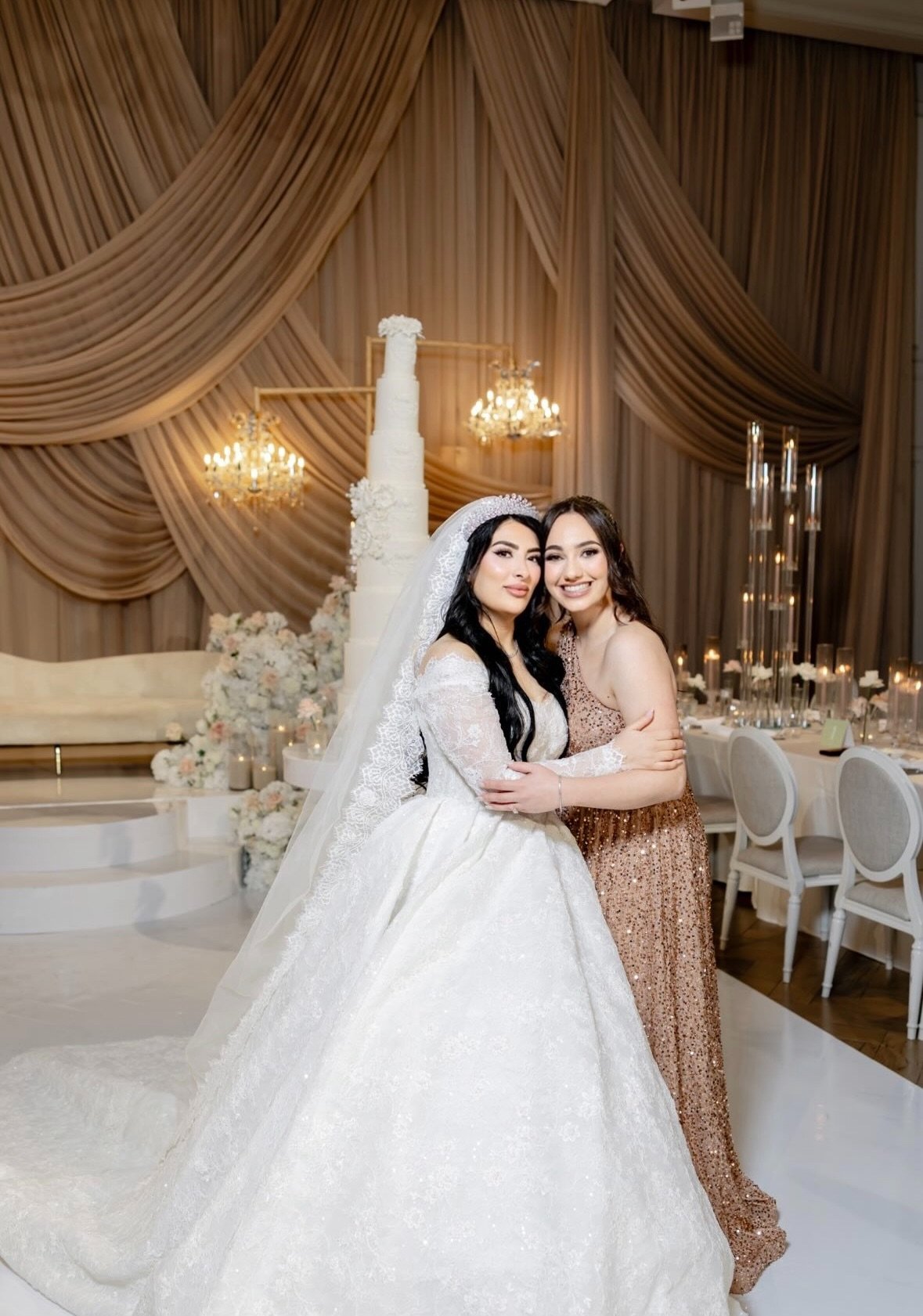 Two women, one in a wedding gown and veil, and the other in a sparkling dress, embracing and smiling at a wedding reception decorated with drapes, floral arrangements, candles, and chandeliers.