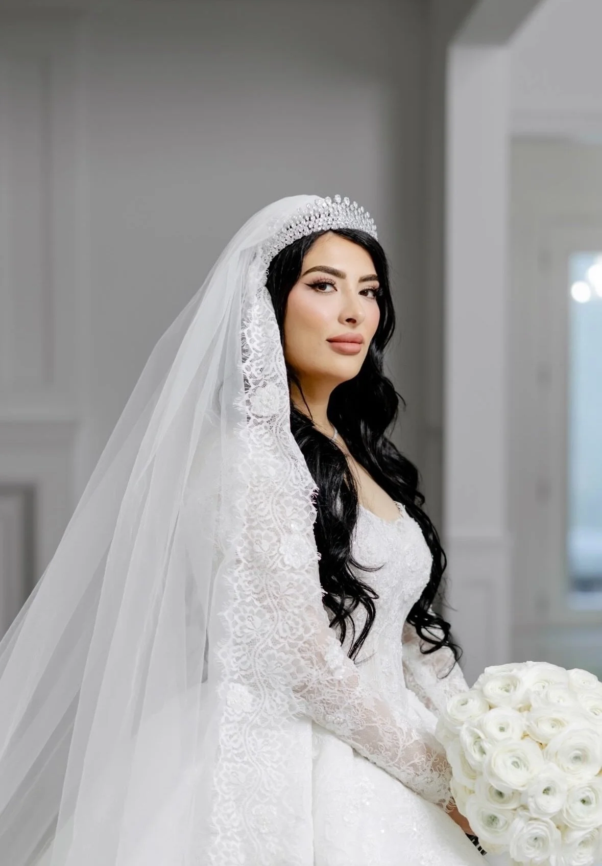 Bride with long black hair in a white lace wedding dress holding a bouquet of white roses, standing in a bright indoor space.