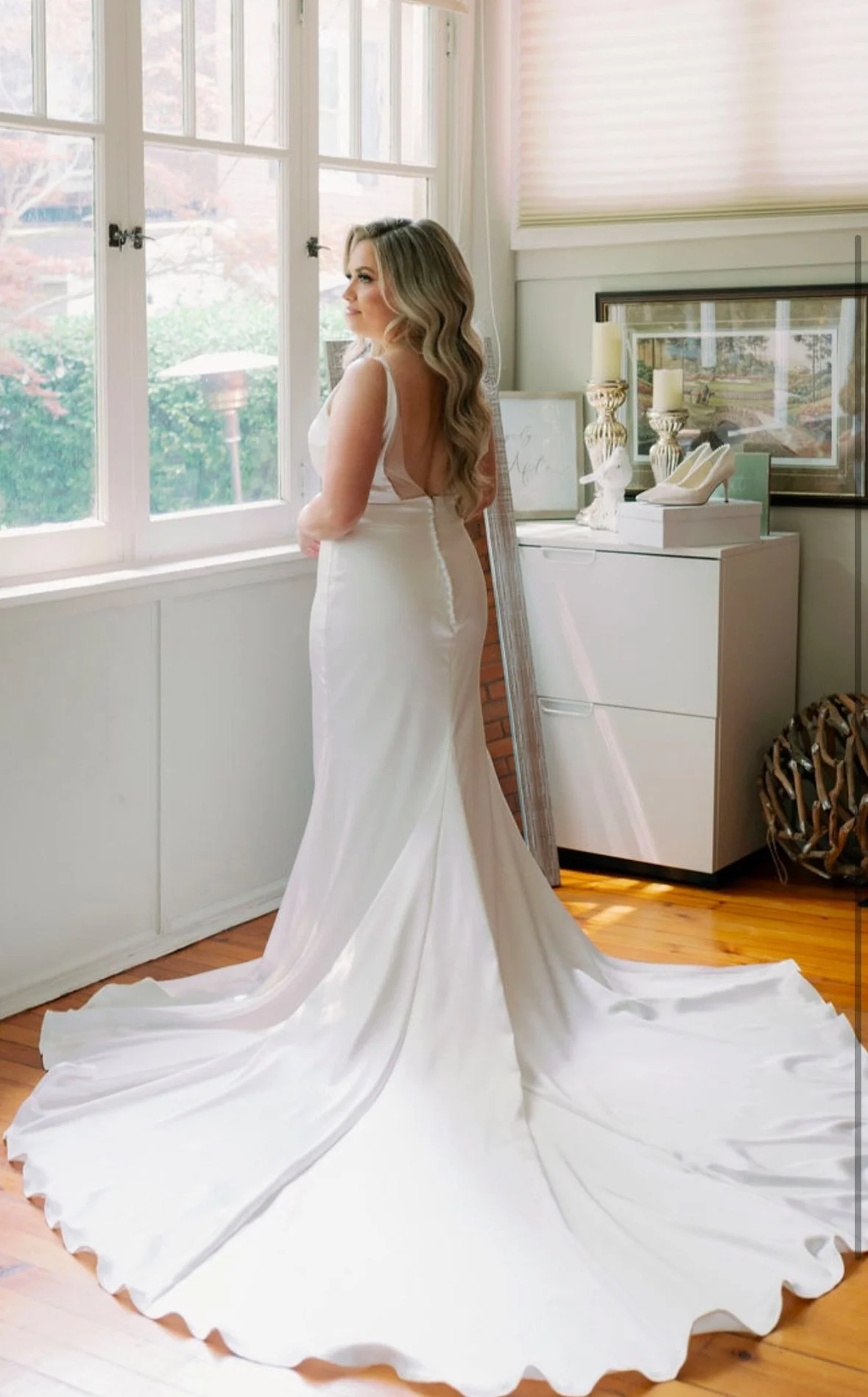 A woman in a white wedding gown standing by a window, with a long train flowing on the floor, looking outside.