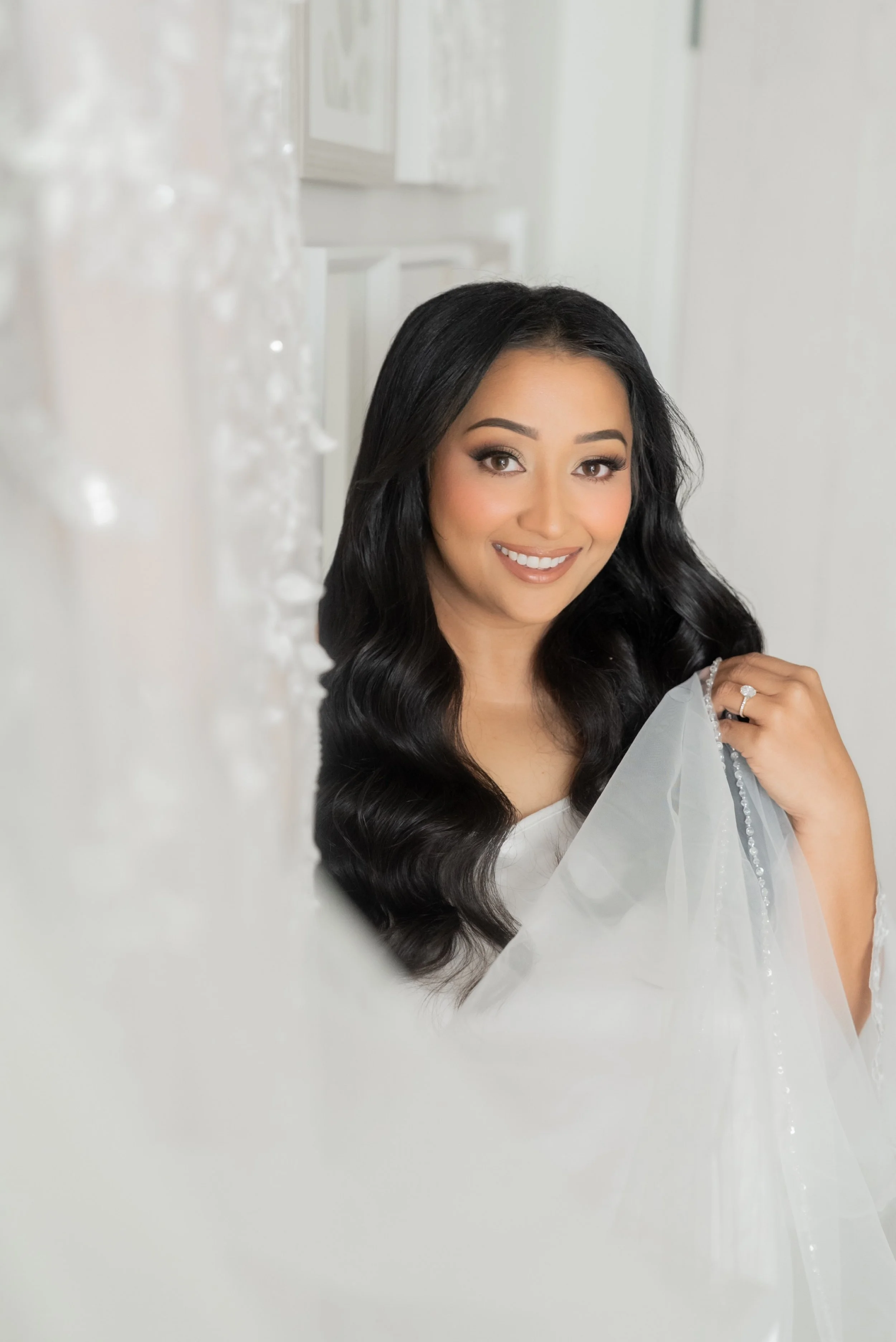 A young woman with long, wavy black hair smiling, holding a sheer white wedding veil, standing indoors near a white wall with framed pictures.