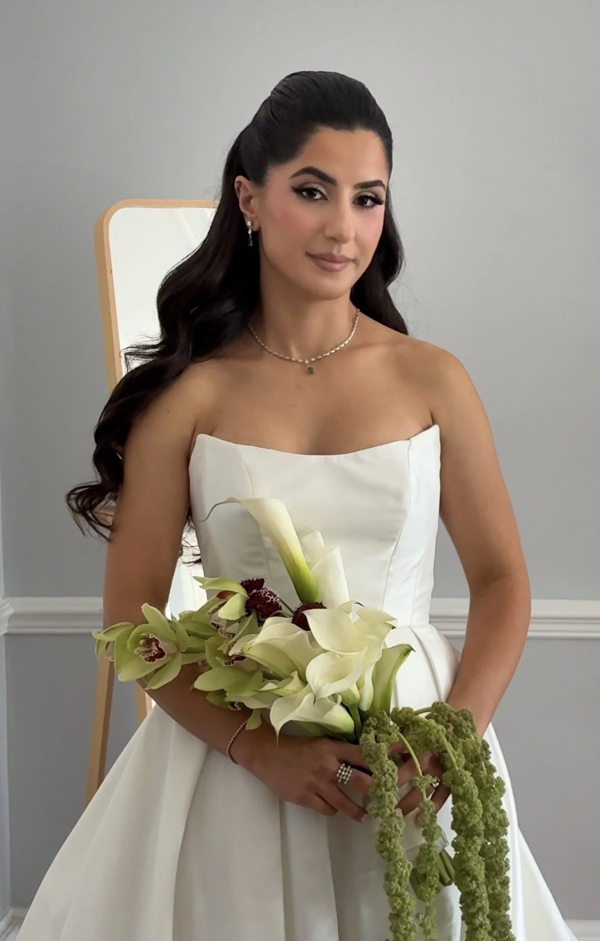 A woman in a white wedding dress holds a bouquet of white calla lilies and other flowers, standing in front of a light-colored wall and mirror.