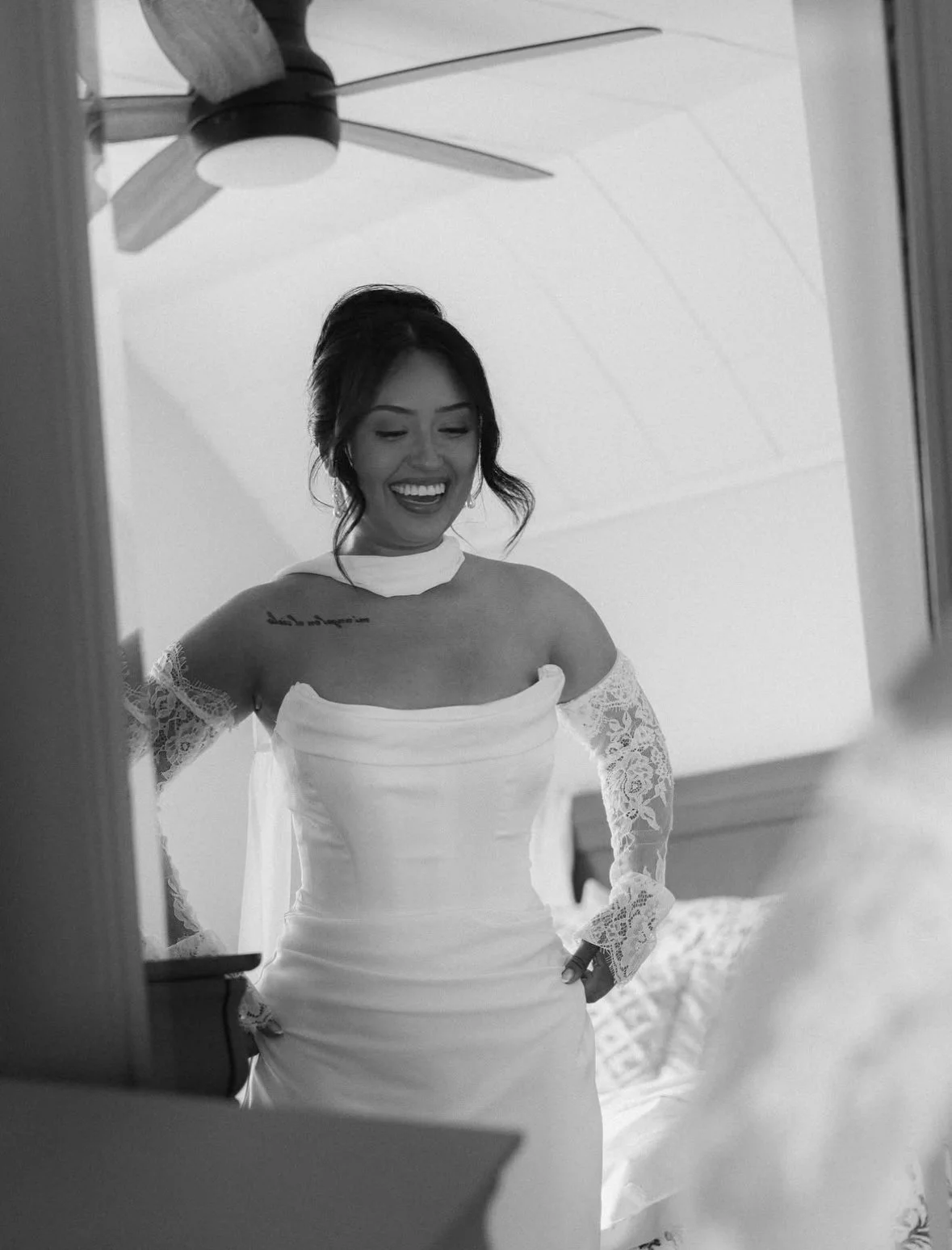 A woman dressed in a wedding gown and lace sleeves, smiling and looking down, standing in a room with a ceiling fan.