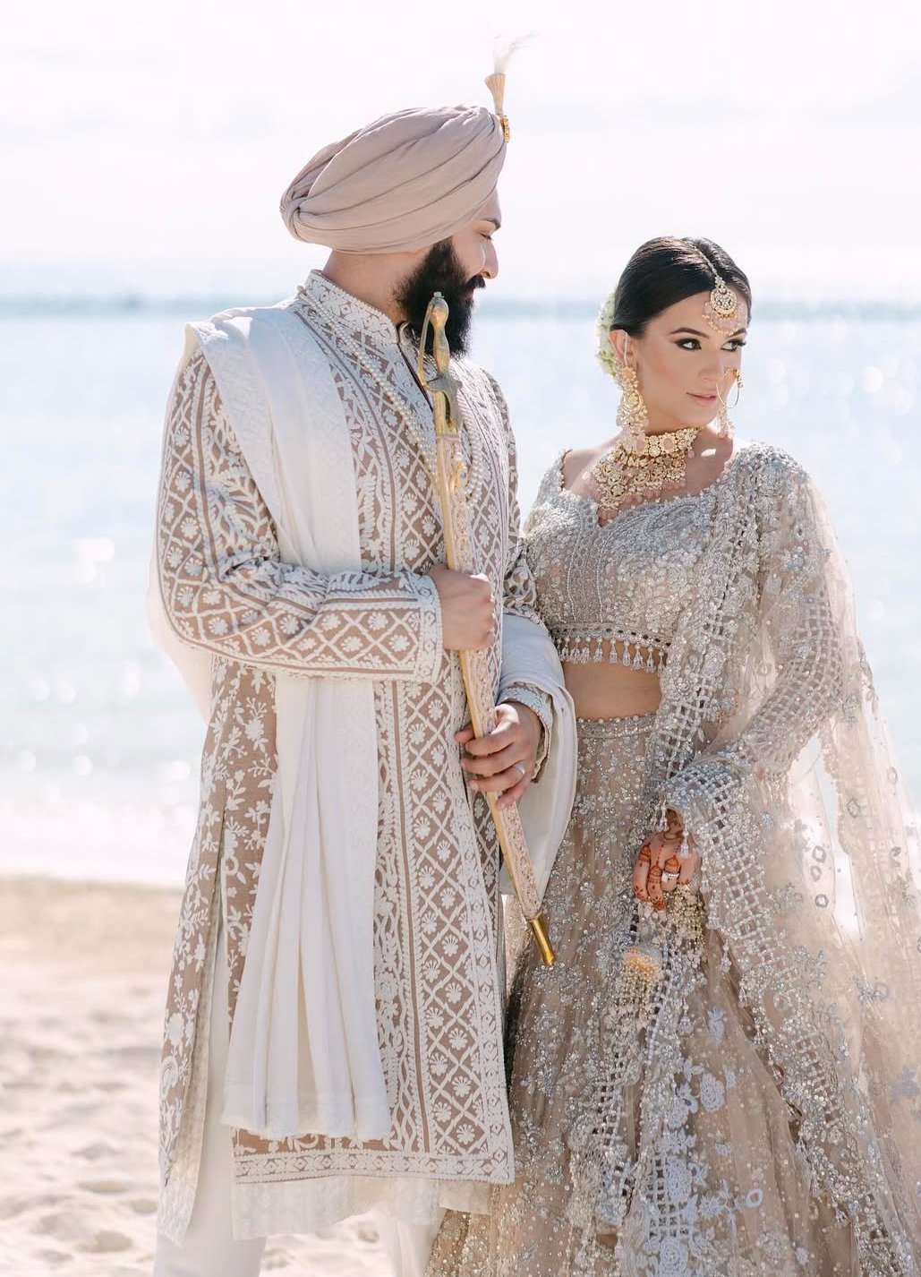 A bride and groom dressed in traditional Indian wedding attire standing on a beach with the ocean in the background.