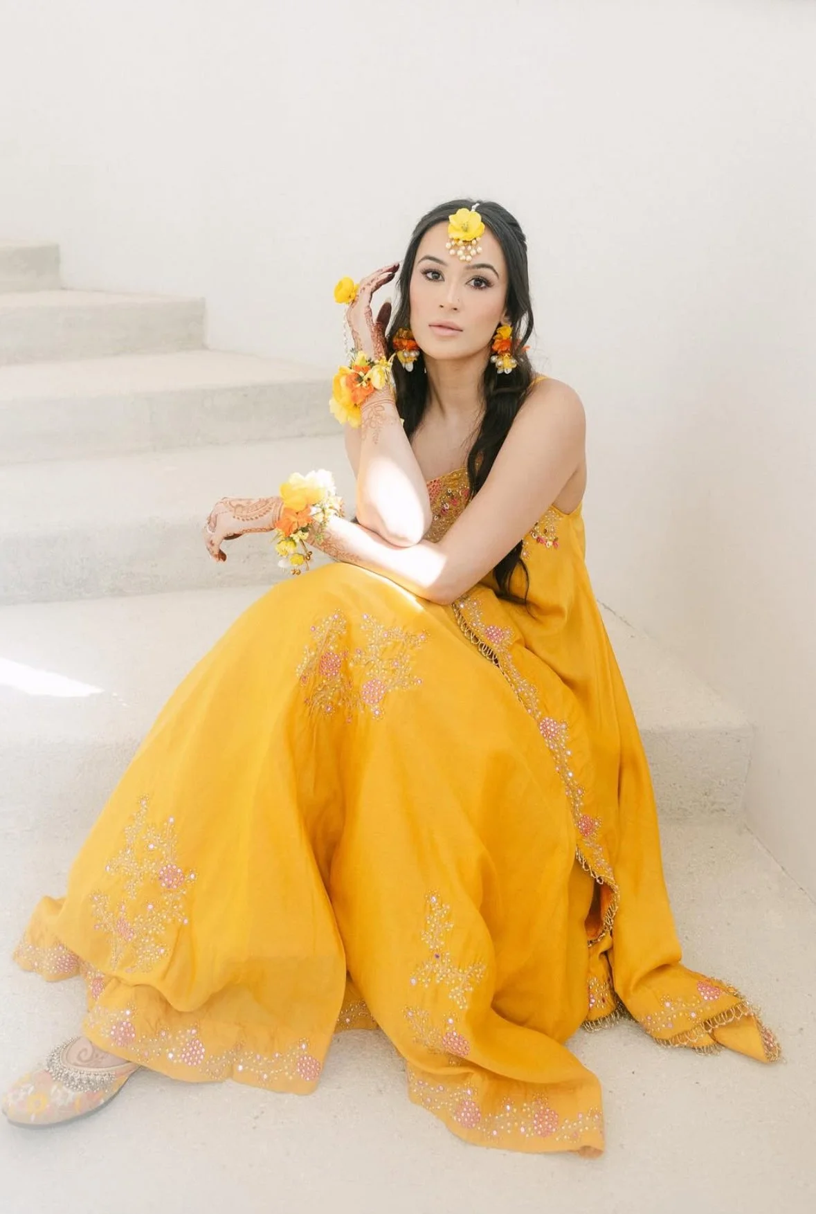 A woman dressed in a bright yellow traditional Indian outfit with embroidery, sitting on a staircase against a plain white wall, wearing floral jewelry and henna on her hands.