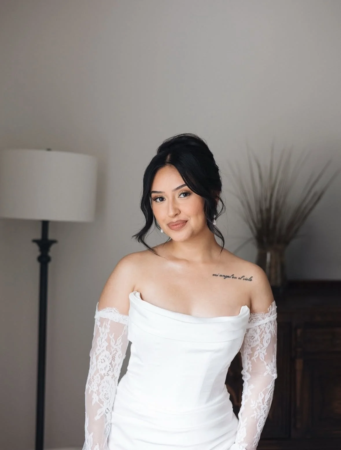 A woman wearing a white off-shoulder dress with lace sleeves, standing in a room with a lamp and a decorative plant in the background.