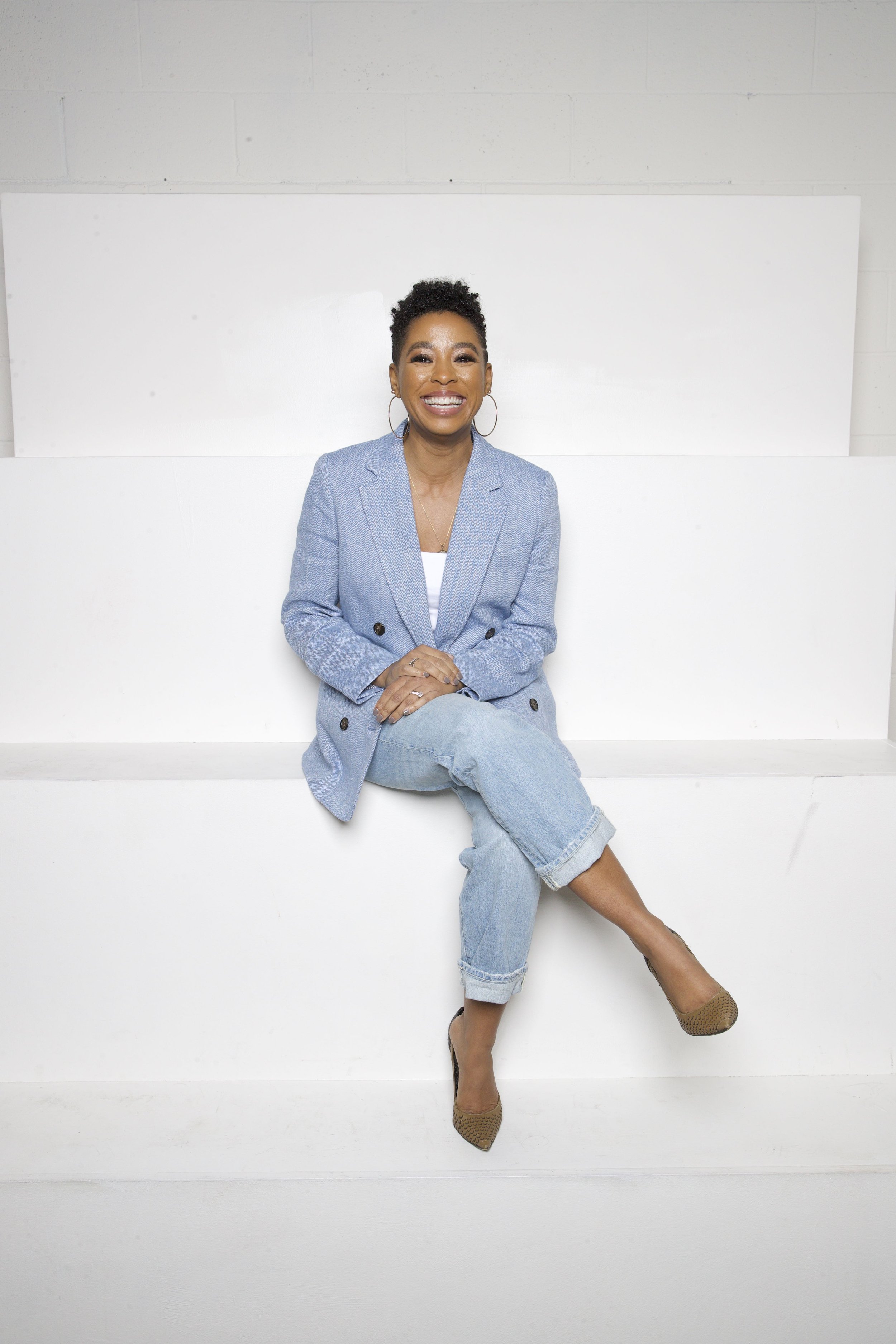 A woman sitting on a white bench, smiling, wearing a light blue blazer, jeans, and high heels, against a plain white background.