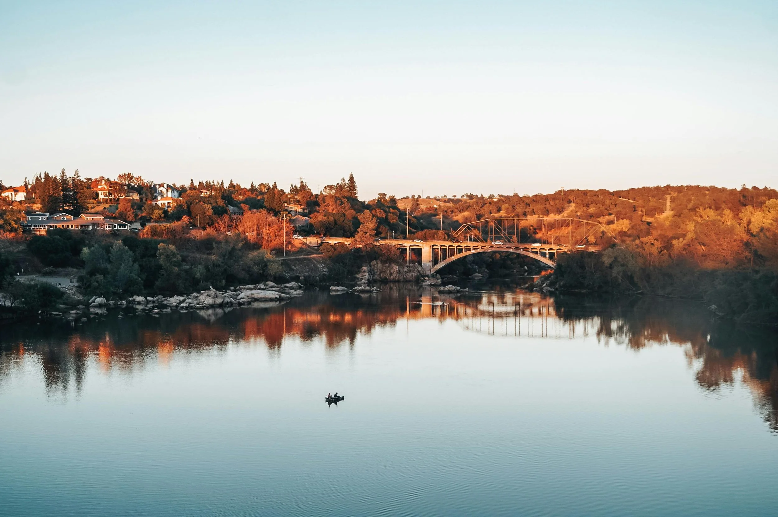 A calm river reflecting the trees and houses on its banks, with a small boat floating on the water, and a bridge spanning the river in the background during sunset.