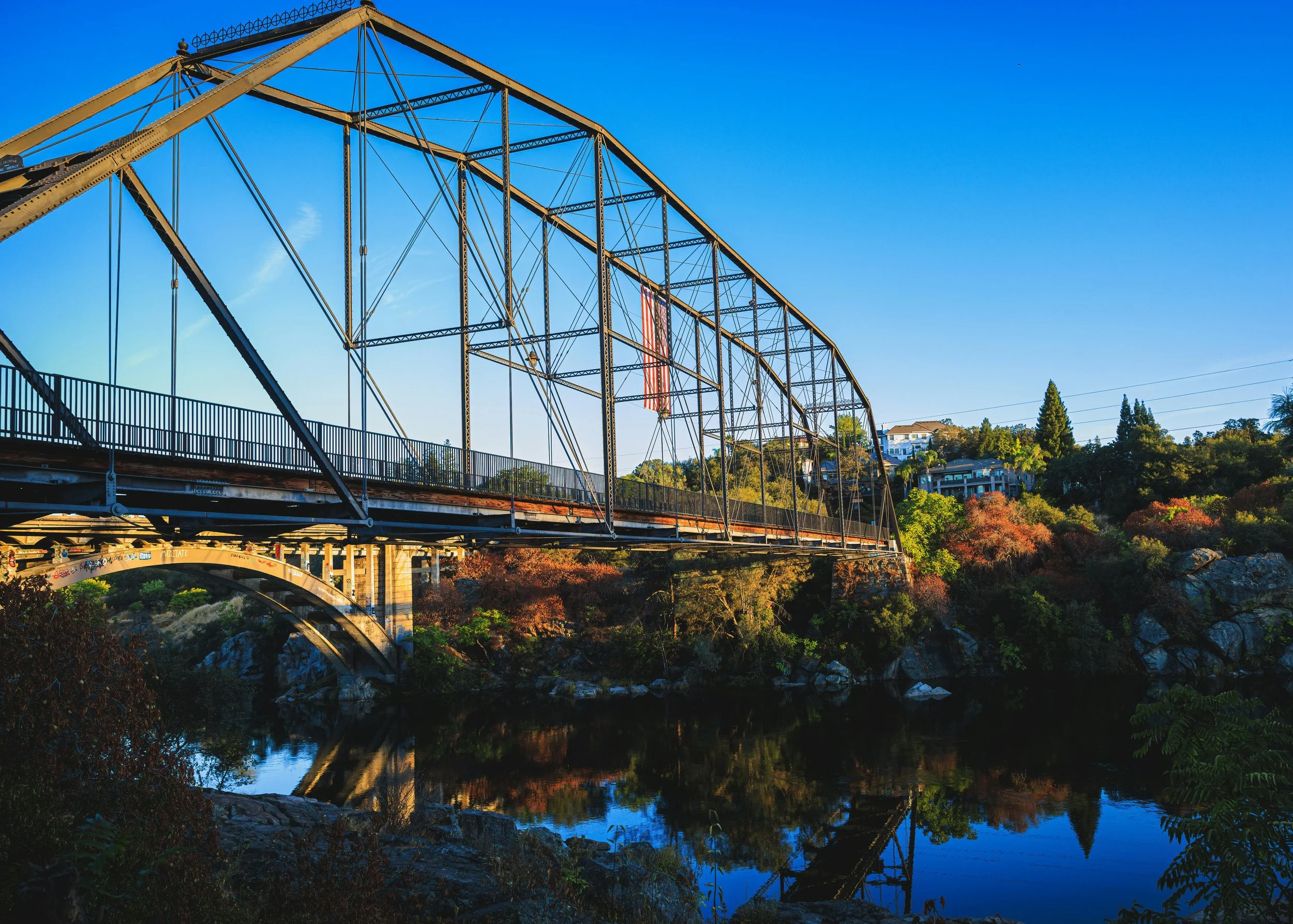 A scenic view of a steel arch bridge over a river, with houses and trees on a hillside in the background under a clear blue sky.