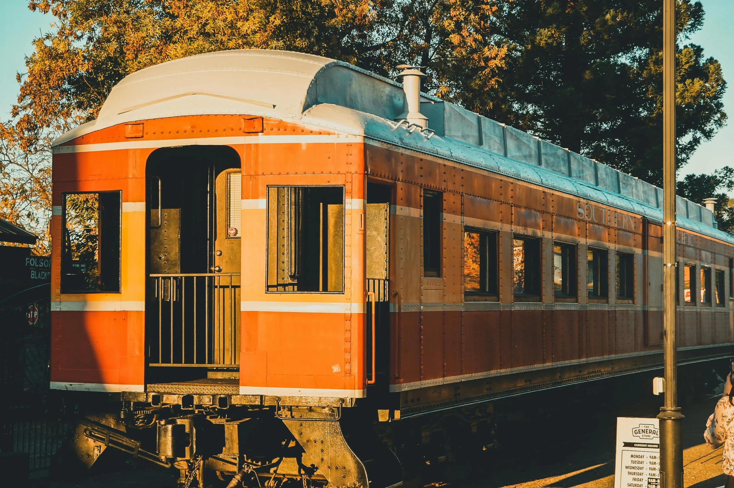 An orange vintage train car with a white roof is parked on the tracks, illuminated by warm sunlight, with trees in the background.