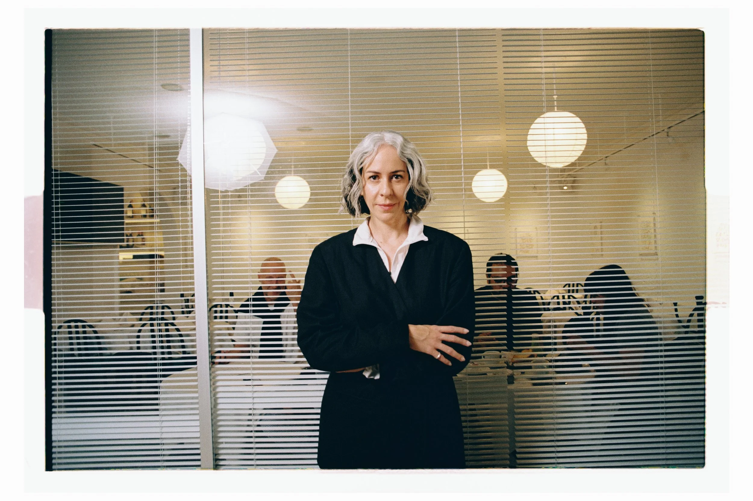 A woman with gray hair and a black blazer standing with arms crossed in front of a glass partition in a room with hanging round paper lantern lights.
