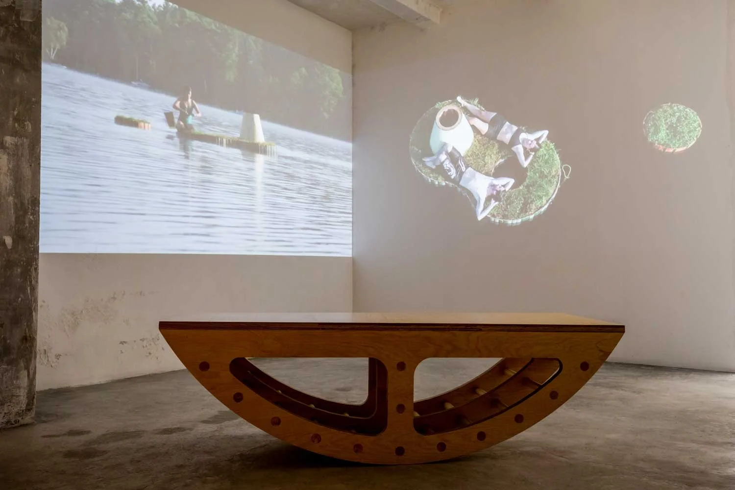 Projection of a woman kayaking on water and two sculptures of people kayaking on grass on wall