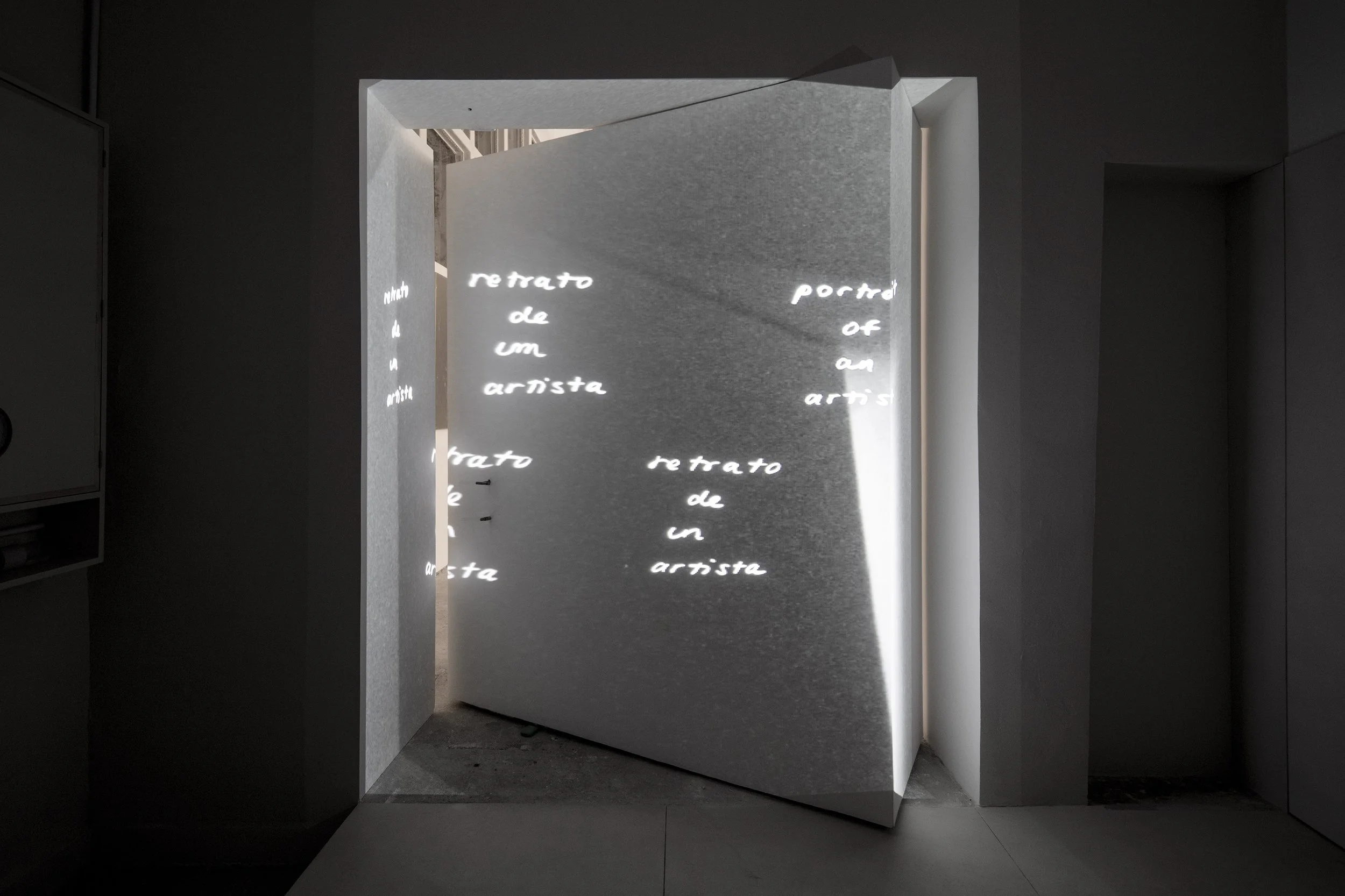 An exhibition space showing a vertical, rectangular display with illuminated text in multiple languages, including Italian and English, on its reflective surfaces. The space has dark surrounding walls and a bright, focused light on the display.