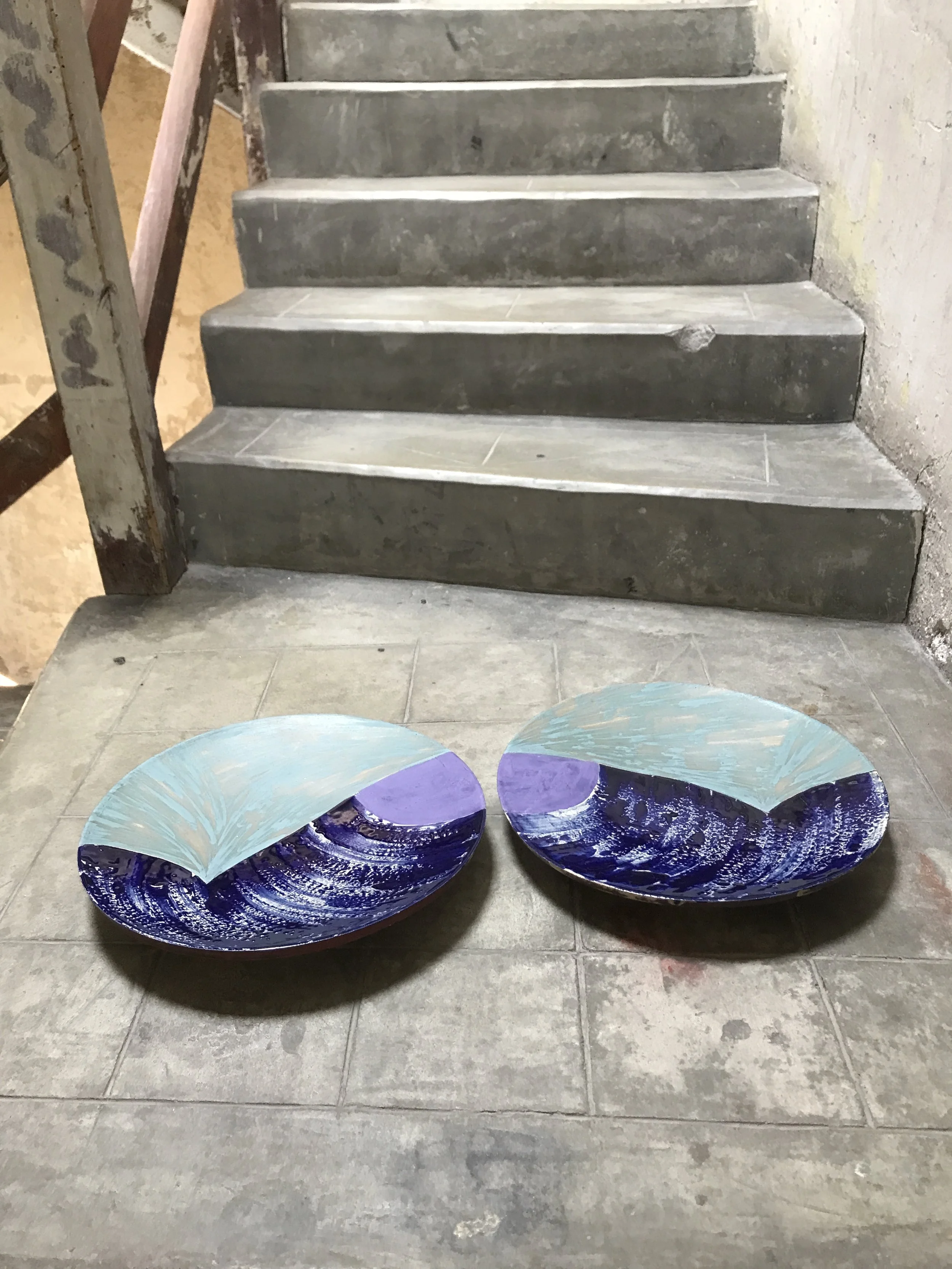 Two decorative glass bowls with blue and purple swirls and a curved glass piece resting inside each bowl, placed on a concrete floor in front of a staircase.