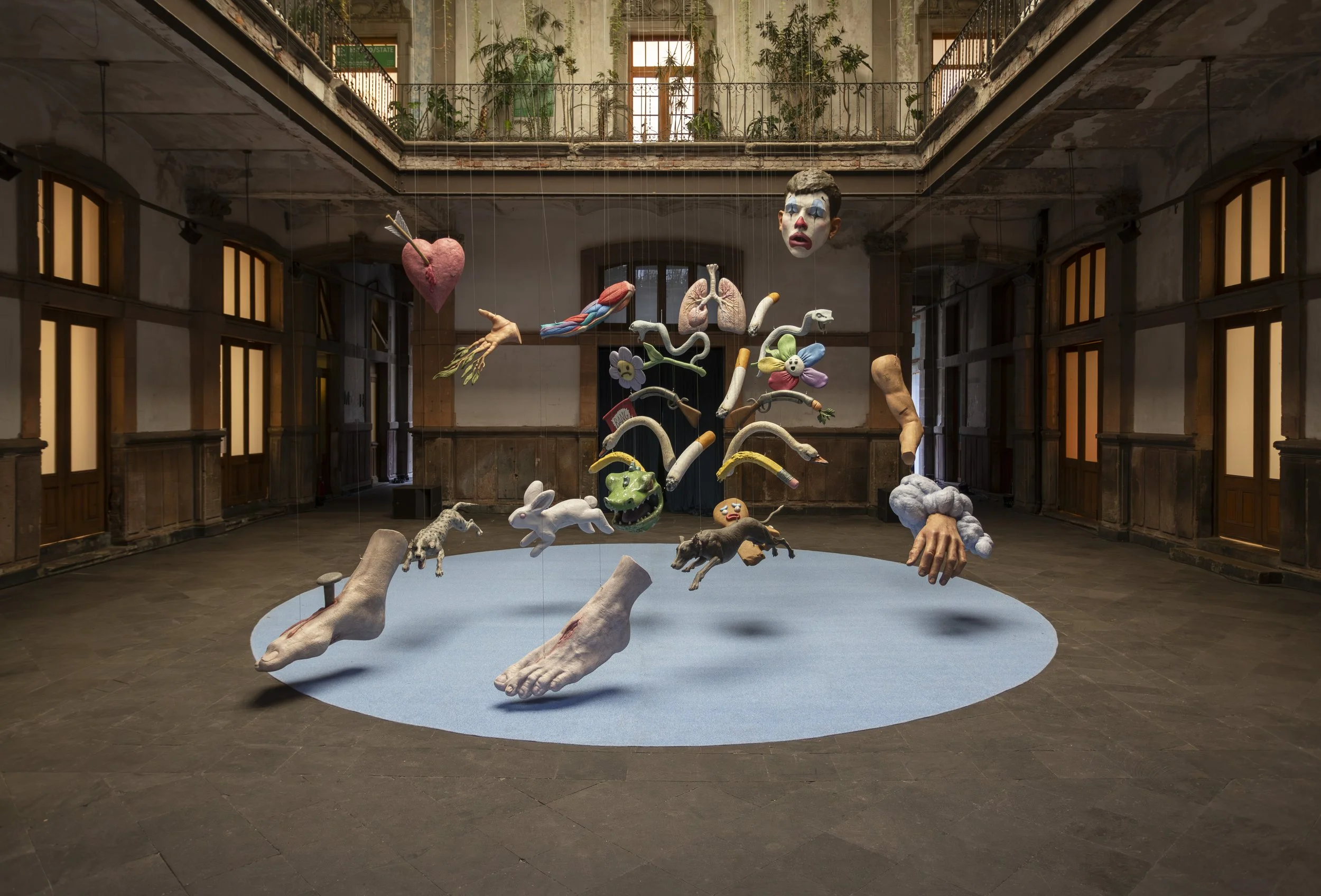 An art installation of suspended mixed media sculptures including human features, animals, and symbolic objects, in an abandoned building with balconies and plants.