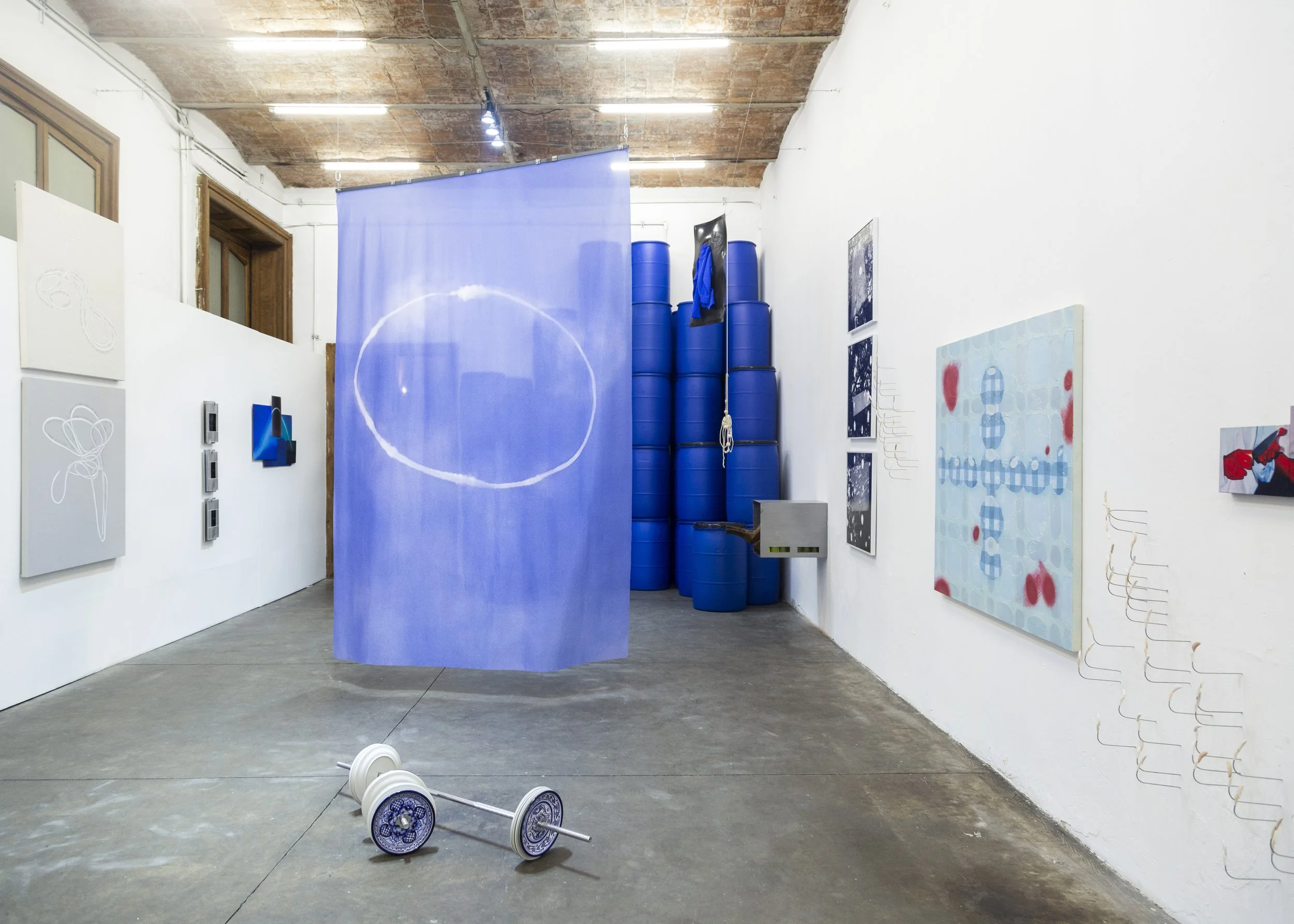 An art gallery with white walls displaying paintings and a large blue curtain or canvas in the center. Blue barrels are stacked in the corner, and a decorative rope hangs on the wall. There are spotlights on the ceiling and some small artworks on the platform in the foreground. A pair of roller skates with decorative wheels is on the gray concrete floor.