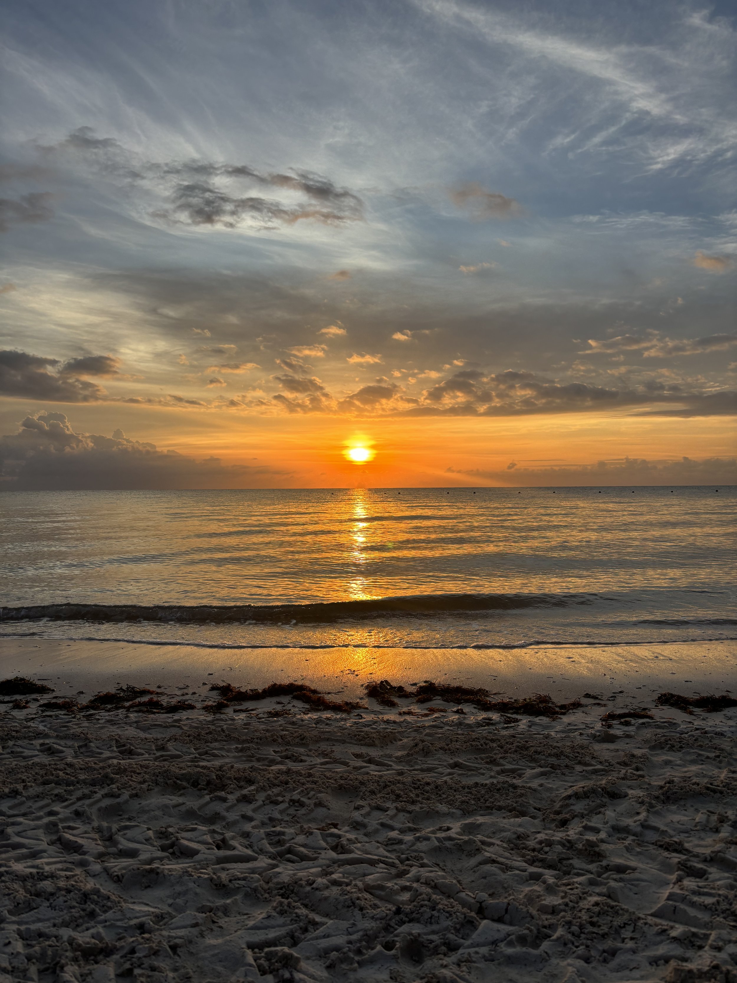 Sunset over the ocean with a partly cloudy sky and a sandy beach in the foreground.