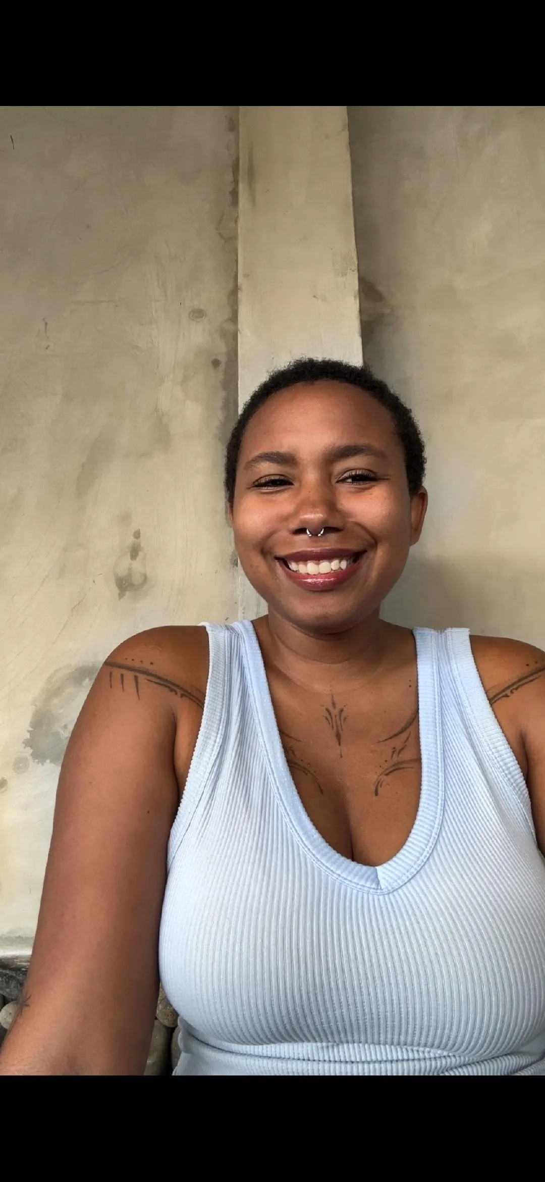 A smiling person with short hair and tattoos on arms and chest is taking a selfie in front of a plain, slightly textured beige wall. They're wearing a light blue ribbed tank top and have a septum piercing.