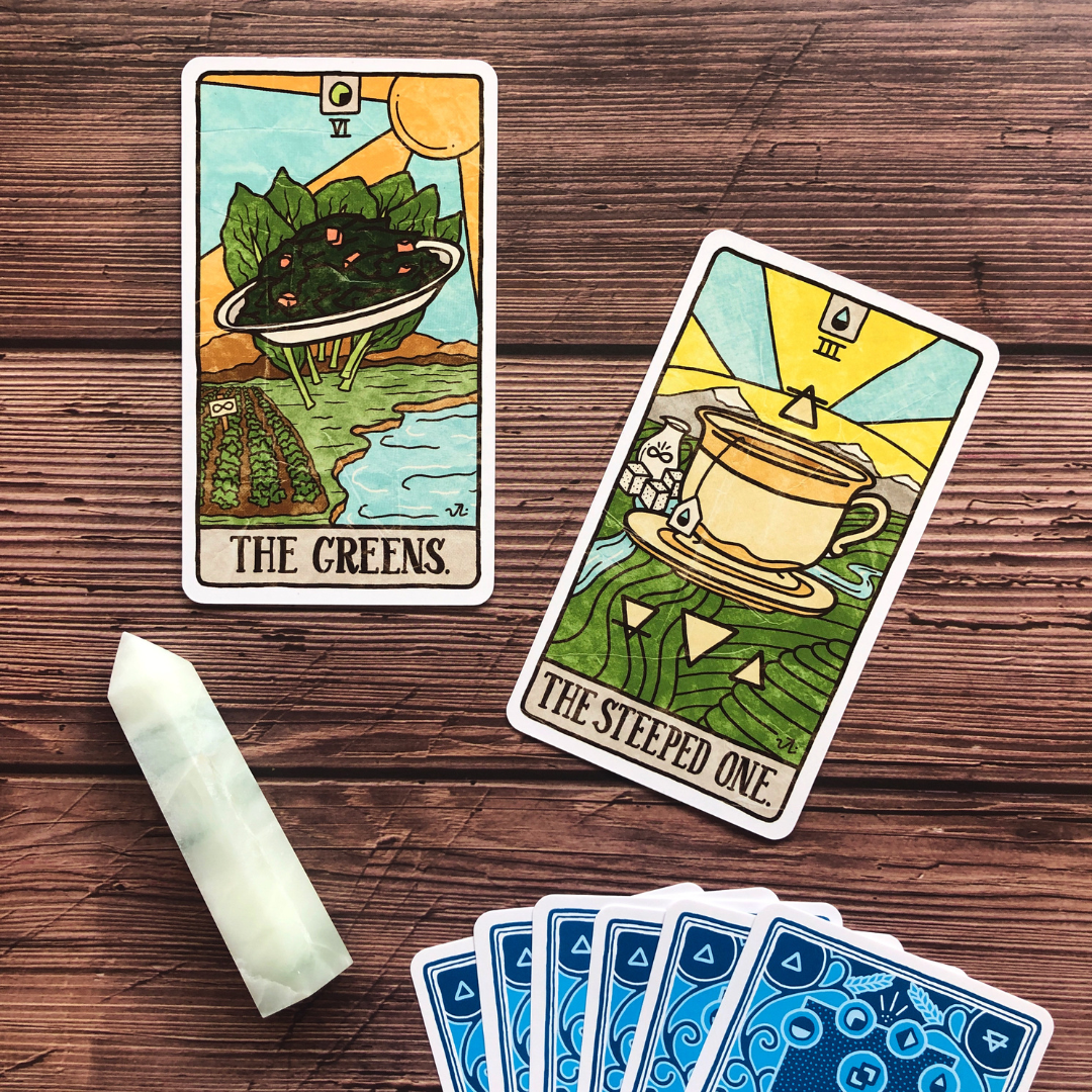 Two illustrated tarot cards, 'The Greens' showing a landscape with leaves and a river, and 'The Steeped One' depicting a teacup with sunlight overhead, lying on a wooden surface alongside a white stone and a spread of blue patterned cards.
