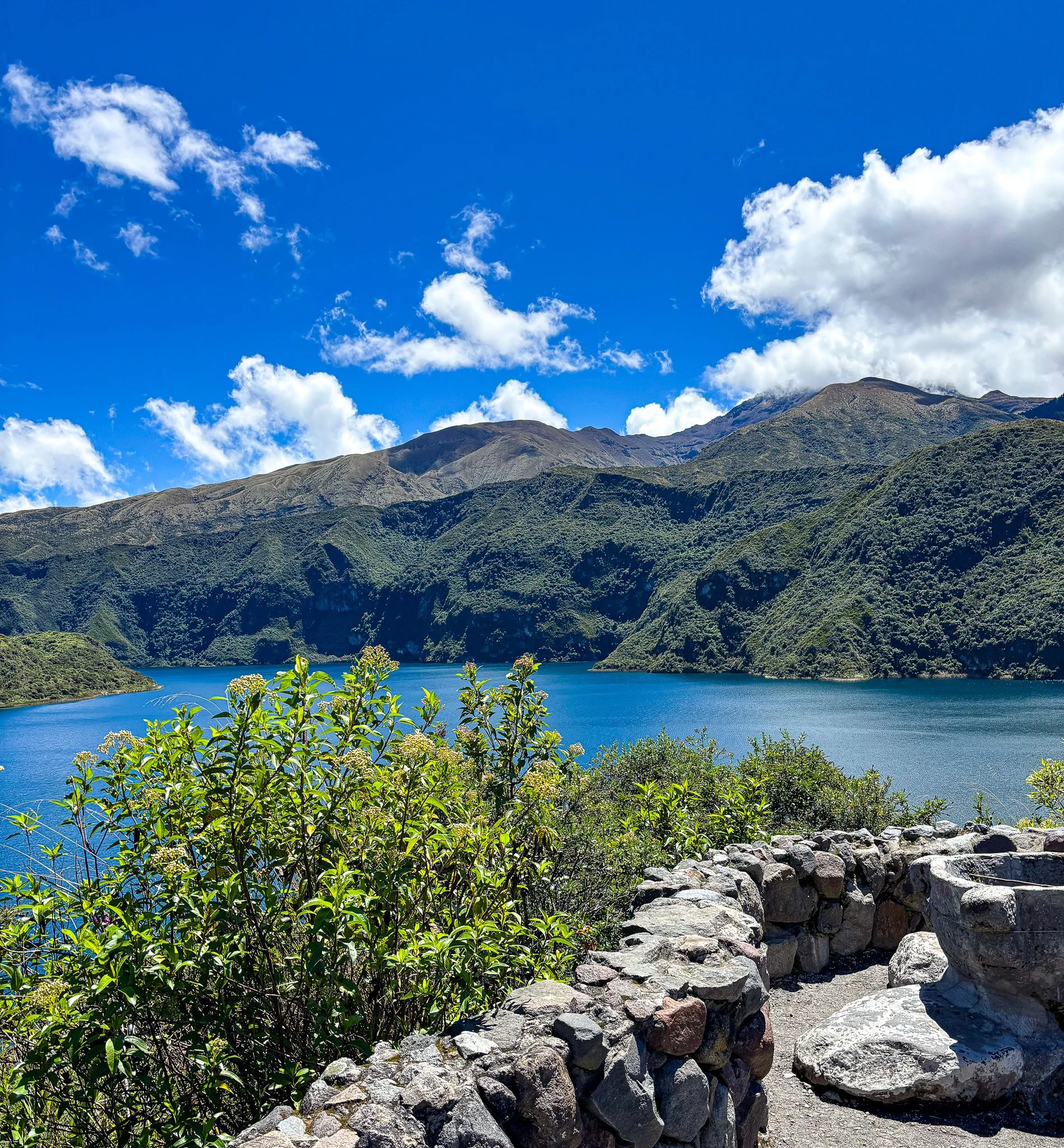 A scenic mountain landscape with lush green slopes, a bright blue lake in the foreground, and a clear sky with scattered white clouds.