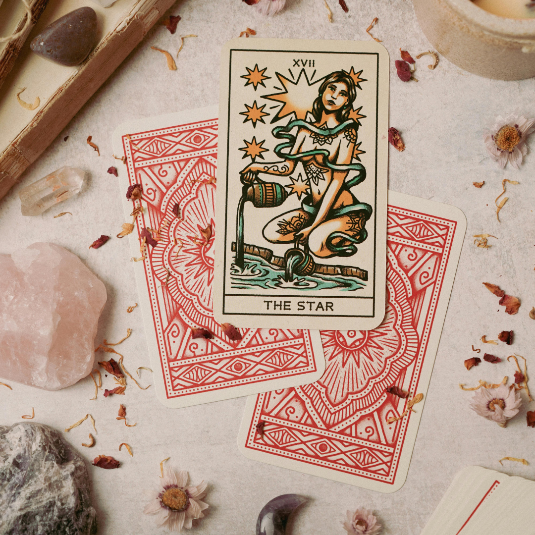 A tarot card called 'The Star' is placed on a surface surrounded by scattered flowers, rocks, and crystals. The card depicts a woman kneeling by water, pouring water from two jugs, with stars shining above her.