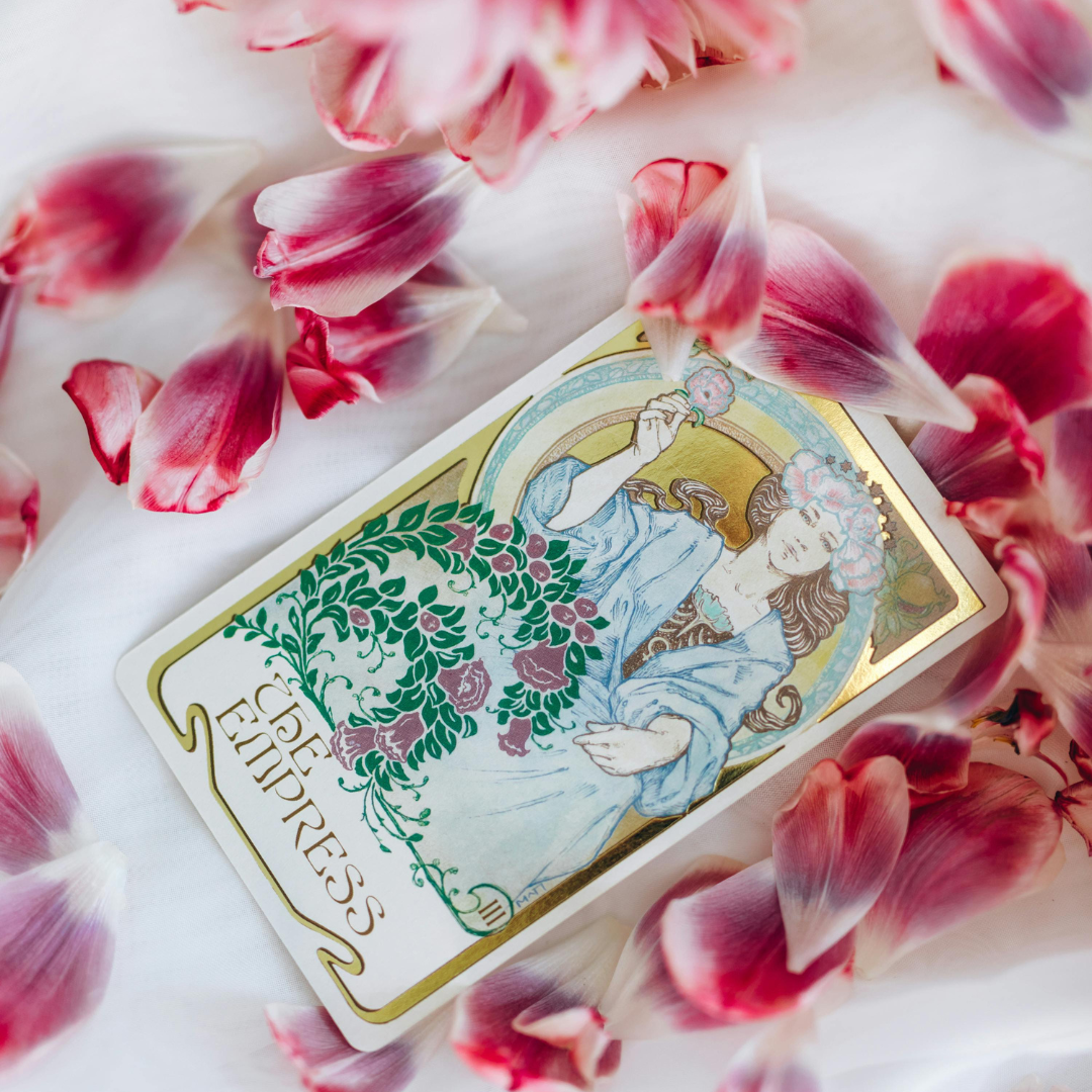 A tarot card titled 'The Empress' surrounded by pink and white flower petals on a white surface.