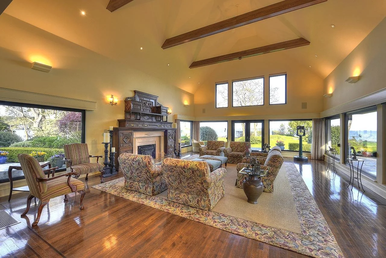 Spacious living room with large windows, wood ceiling beams, vintage furniture, fireplace, and hardwood floors, overlooking a lush outdoor garden.