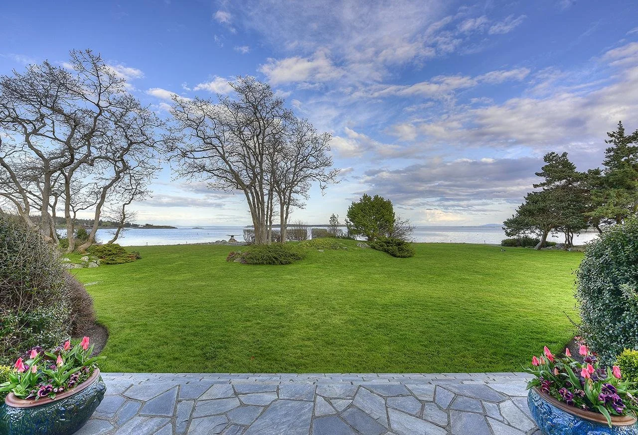 A scenic view of a well-maintained lawn with trees and shrubs, overlooking a body of water under a partly cloudy sky, with two flower pots filled with pink flowers in the foreground.