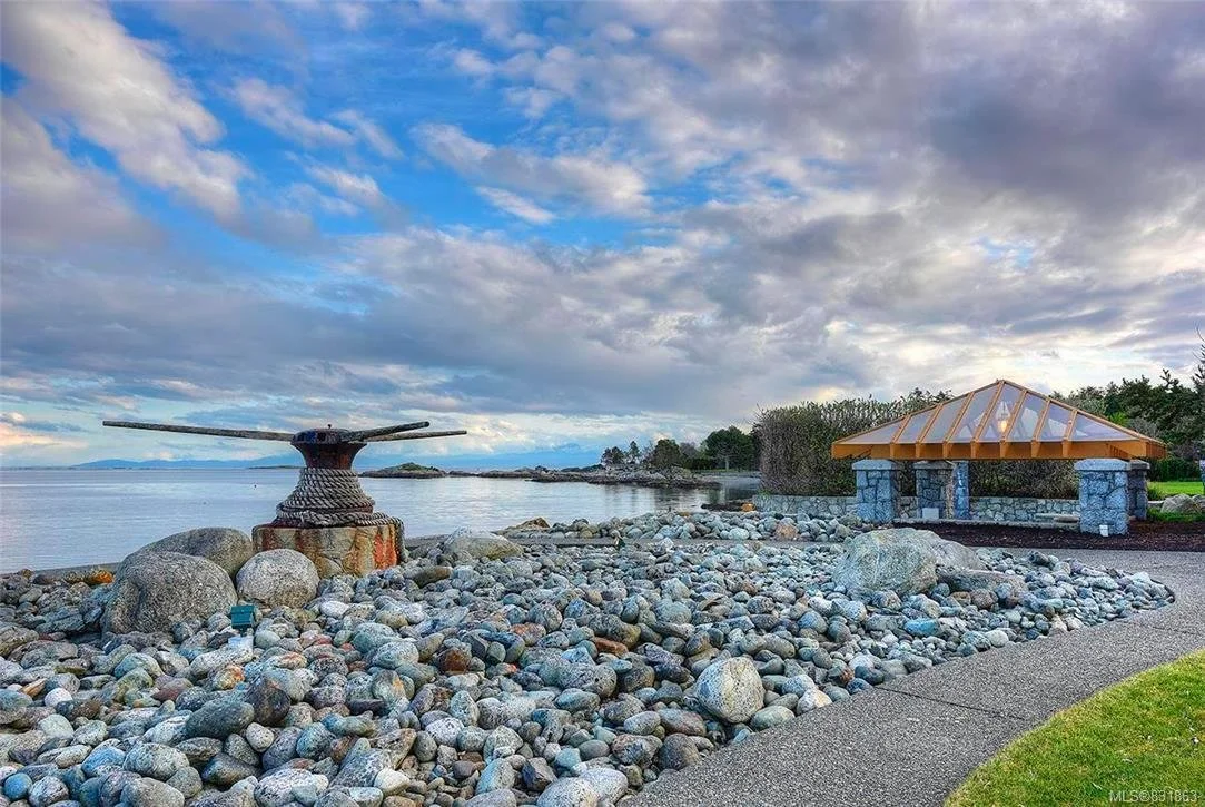 A rocky beach with a large anchor sculpture and a small shelter with a glass roof, overlooking the water and a cloudy sky.
