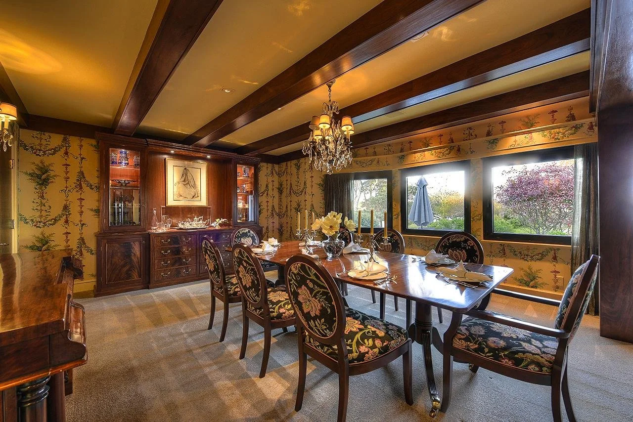 Elegant dining room with a polished wooden table, floral upholstered chairs, a crystal chandelier, and large windows overlooking a garden with trees and an umbrella.