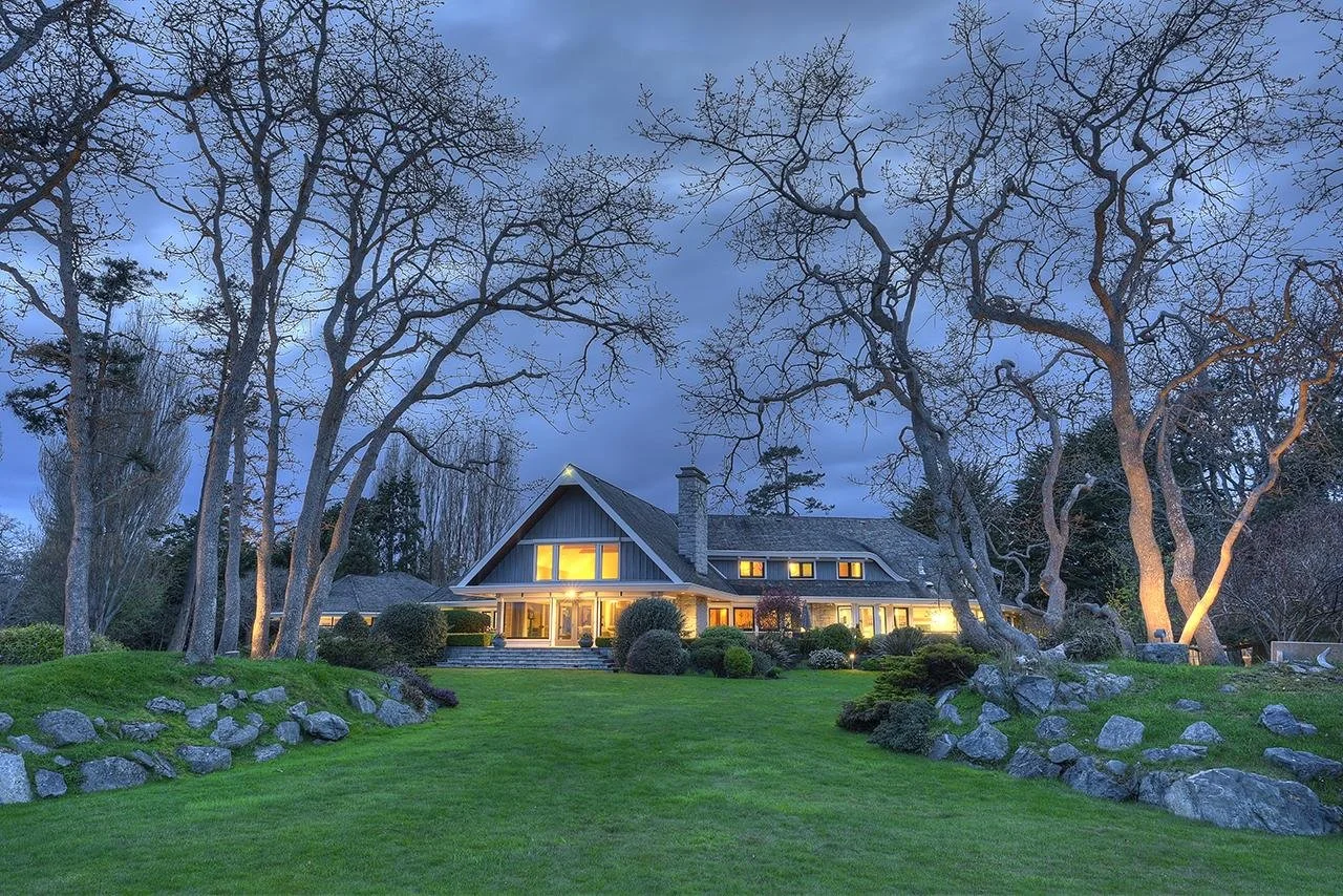 Large house with illuminated windows, surrounded by leafless trees, on a well-maintained grassy lawn during dusk.