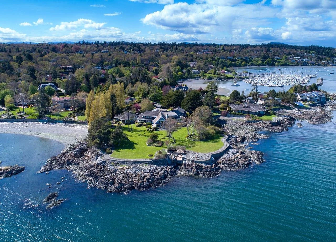 An aerial view of a coastal neighborhood with houses, trees, a rocky shoreline, and a marina with boats in the water.