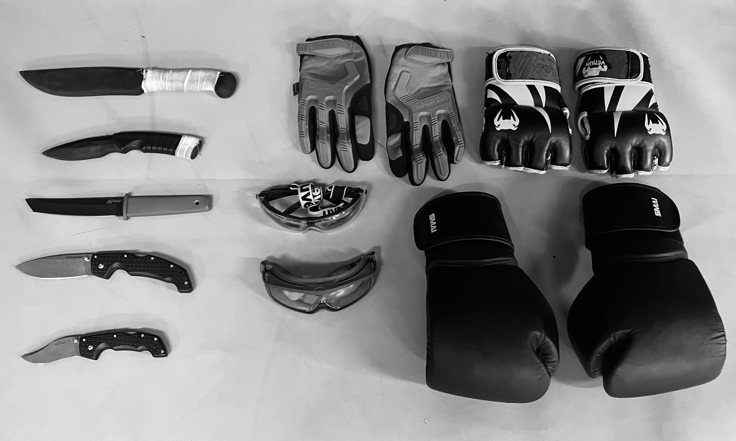 Martial arts or boxing equipment including five knives, two pairs of gloves, a pair of protective headgear, and two pairs of large kicking pads arranged on a flat surface.