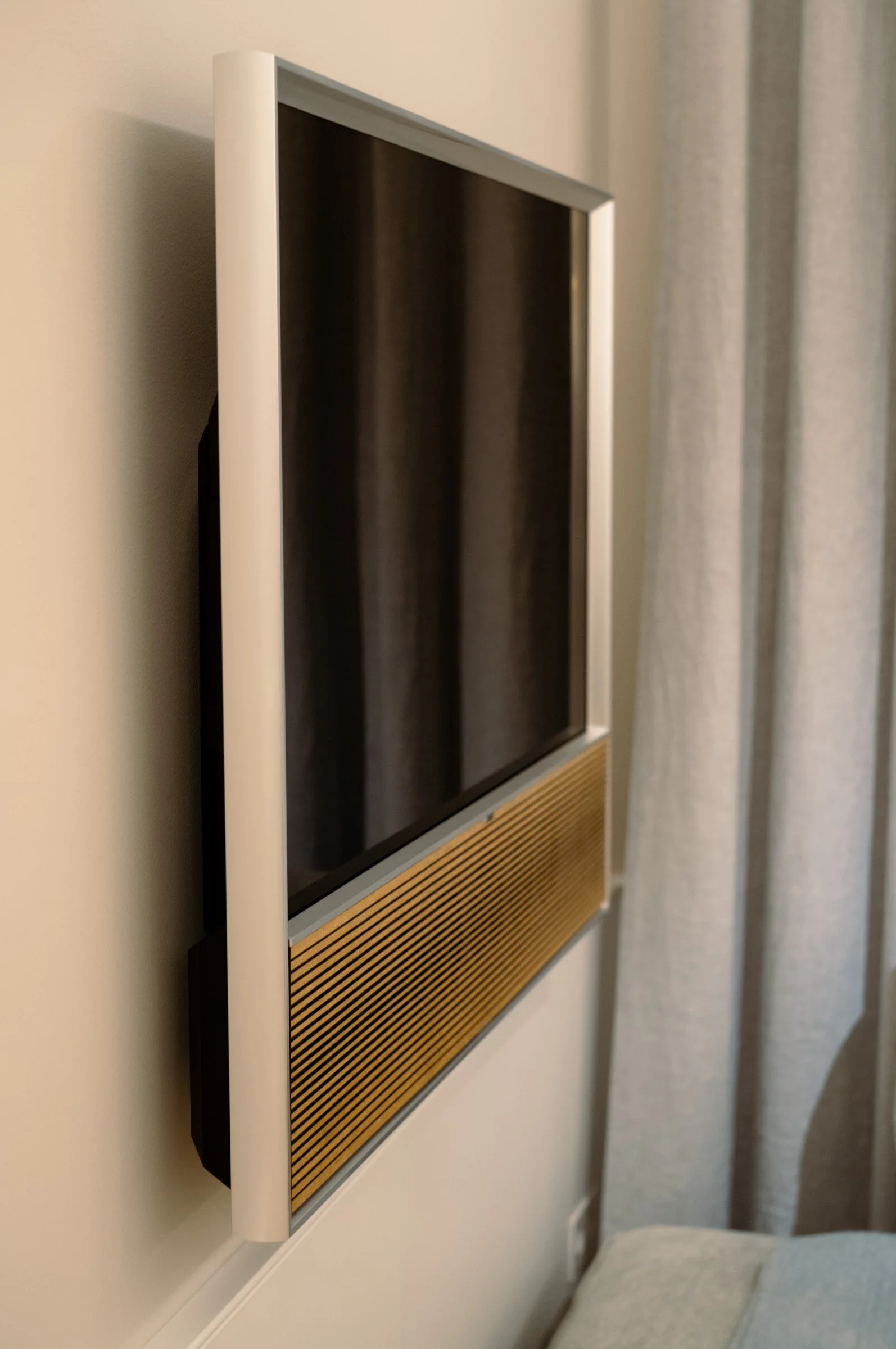 Wall-mounted flat-screen television with a wooden speaker grille below, situated in a modern living space with curtains nearby.