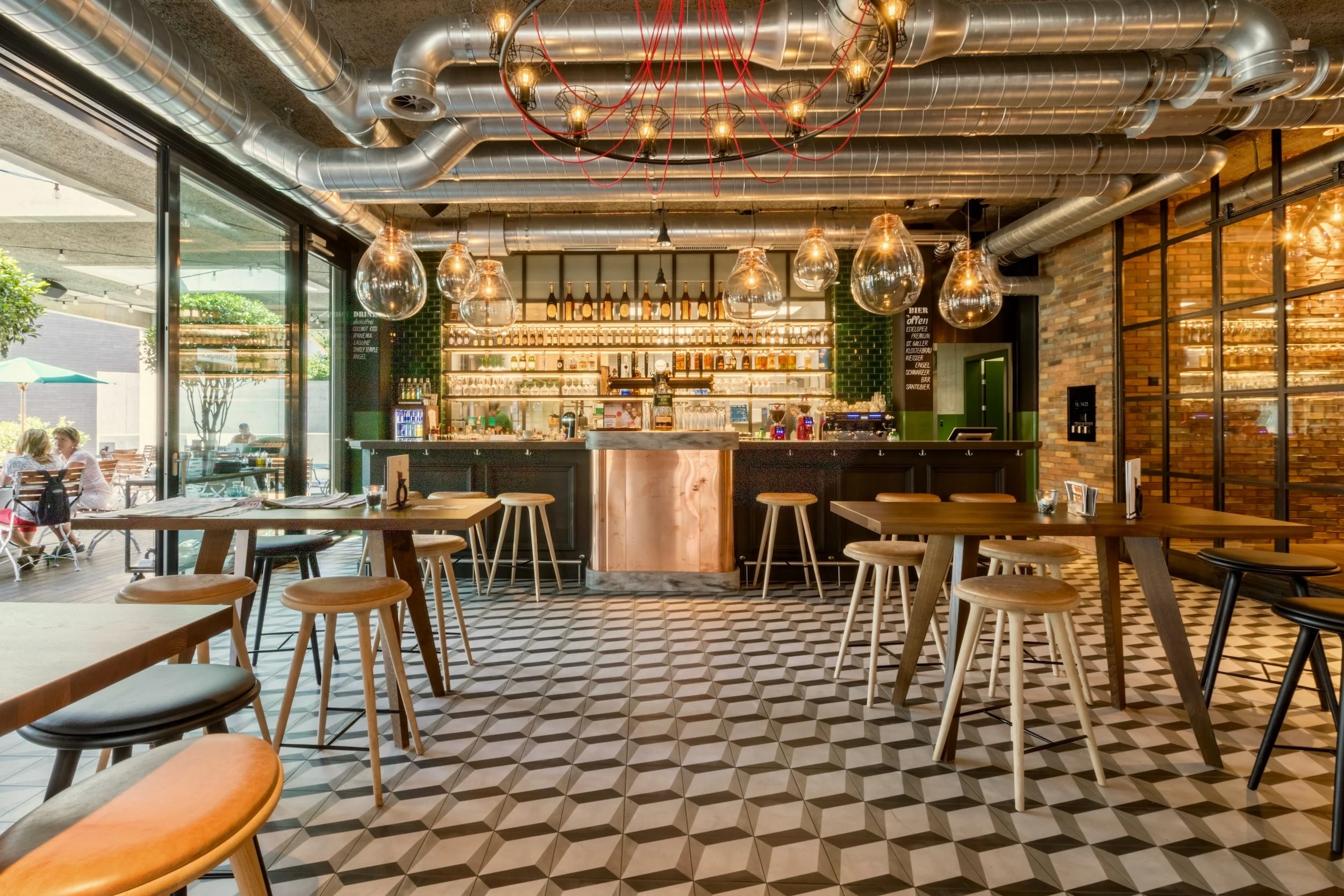 Trendy bar interior with industrial design, featuring geometric tile flooring, wooden tables, stools, and a central bar with hanging light fixtures. Exposed ductwork and shelves stocked with bottles in the background. Outdoor seating visible through large windows.
